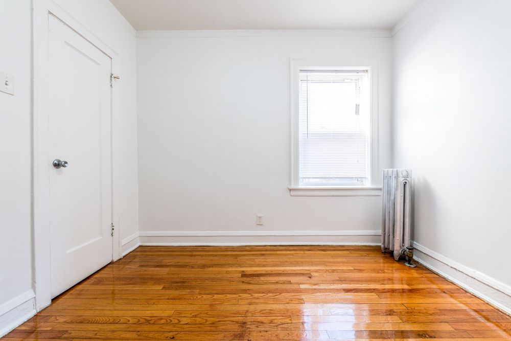 Empty room with hardwood floors, white walls, closet, window, and radiator.