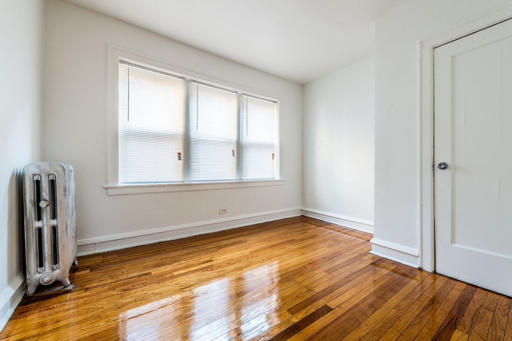 Empty room with hardwood floors, a window with blinds, a radiator, and a white door.