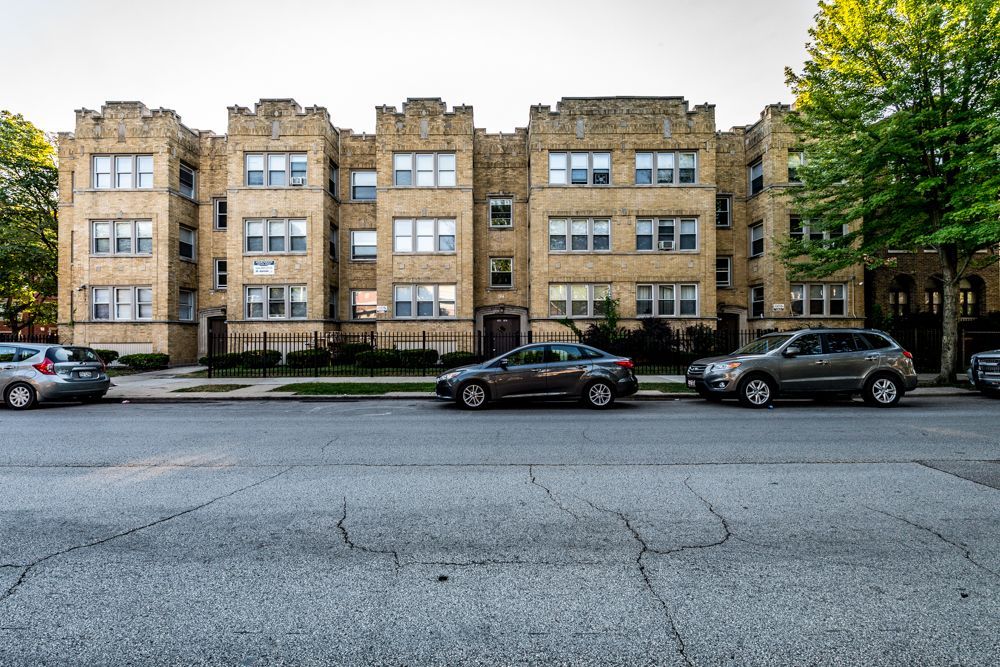 Apartment building with cars parked on the street. Beige brick facade, sidewalk, and trees.