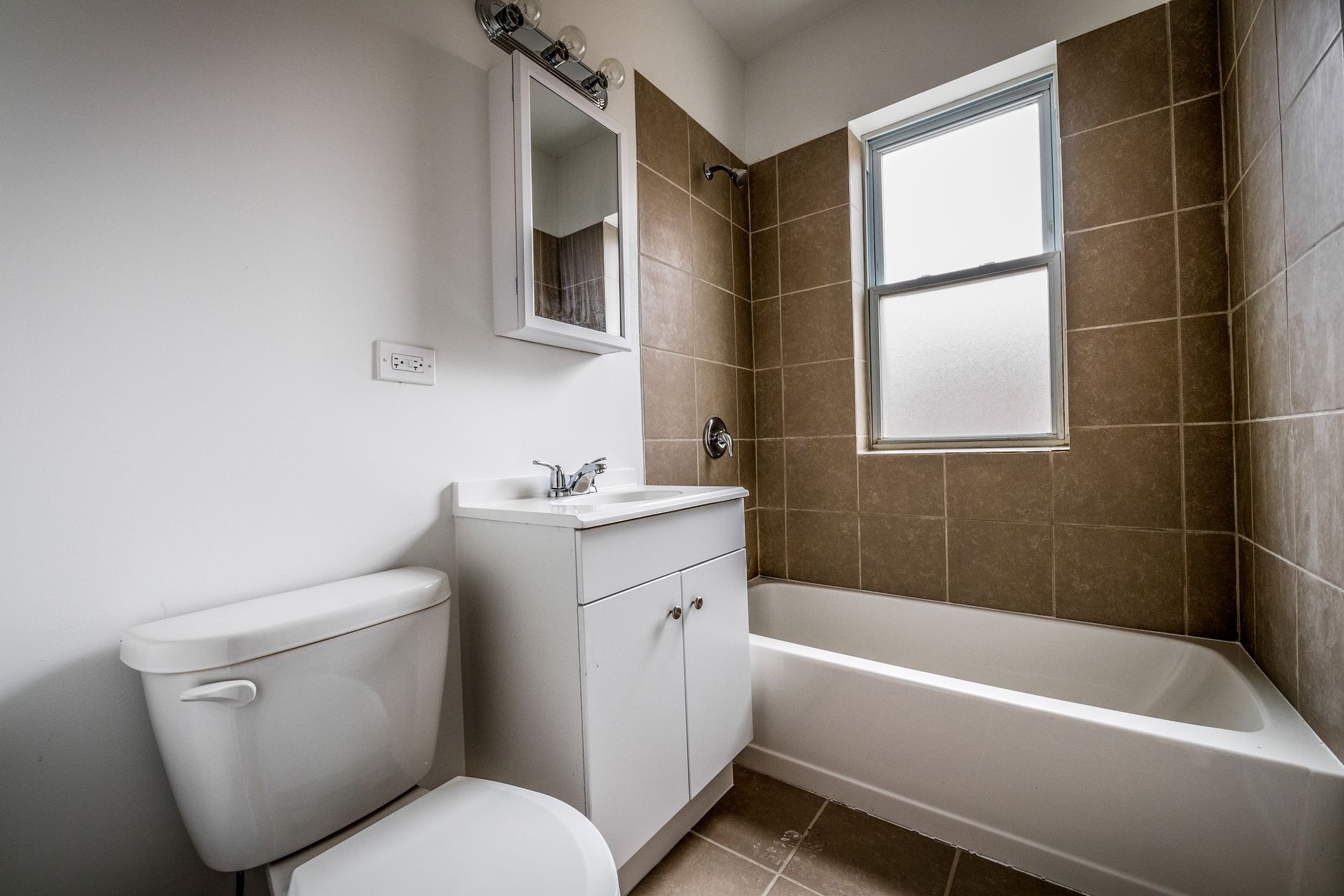 Bathroom with white toilet, vanity, and bathtub. Brown tiled wall, window.