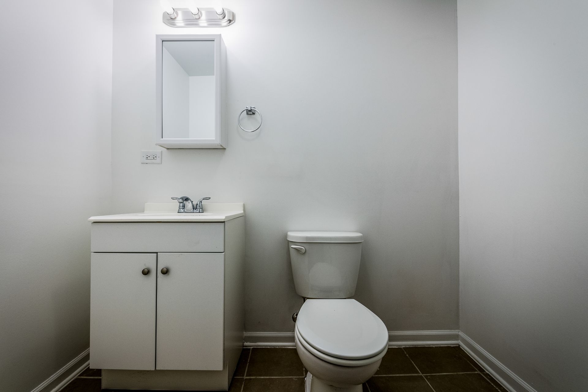 White bathroom with sink, toilet, and cabinet. Brown tile floor, white walls.