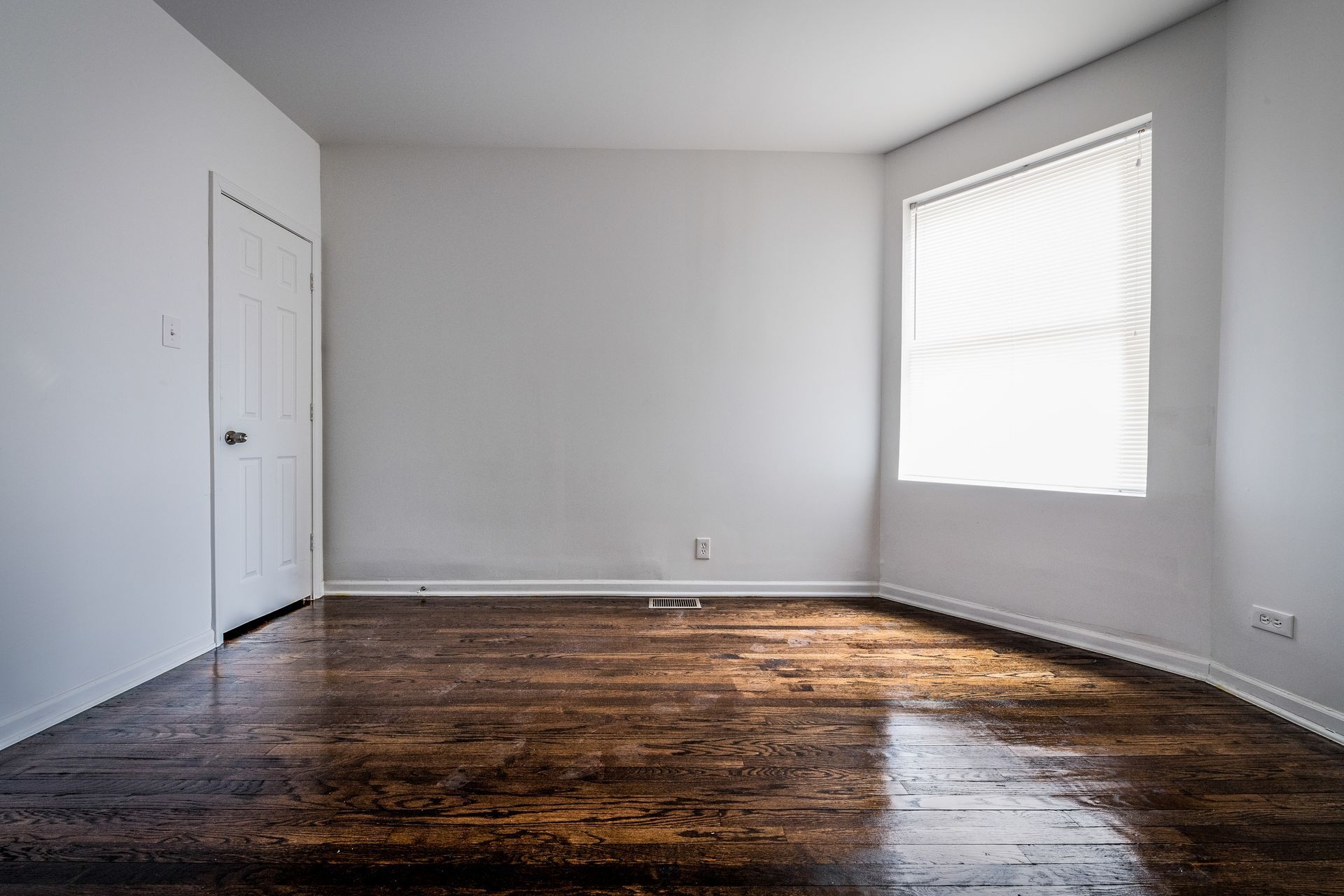 Empty room with dark wood floor, white walls, closed door, and window with blinds.