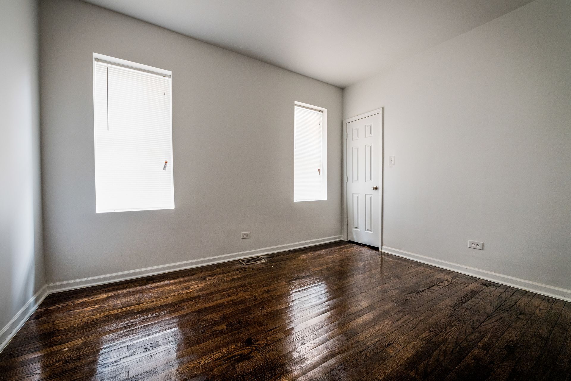 Empty room with wood floor, two windows with blinds, and a closed door.