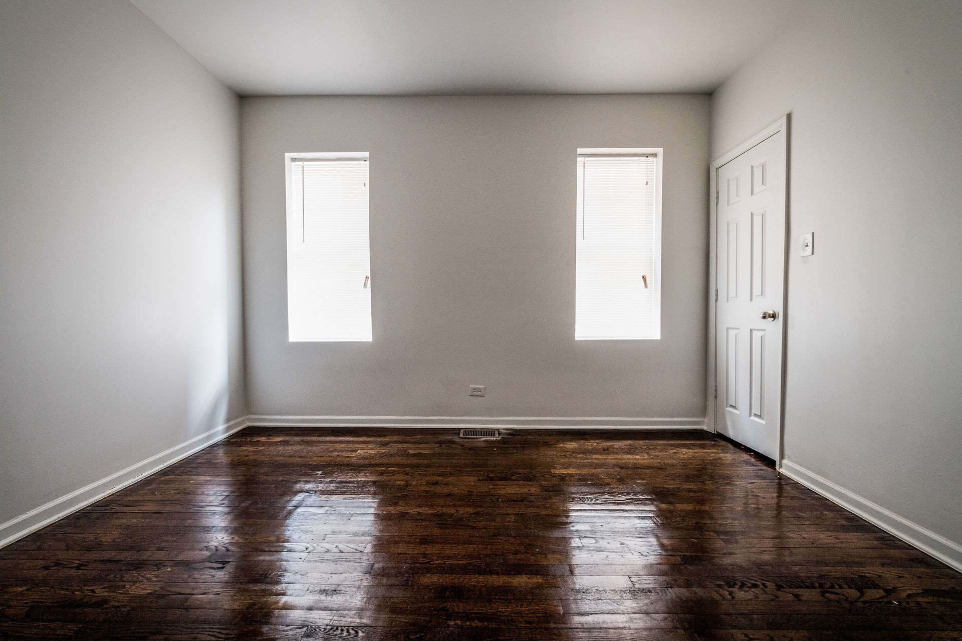 Empty room with wood floors, white walls, two windows with blinds, and a white door.