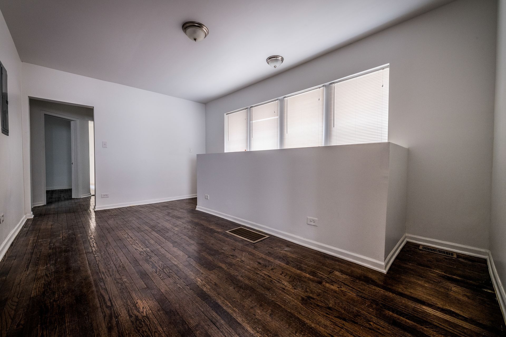 Empty room with wood floors and white walls, small windows with blinds, and a doorway.