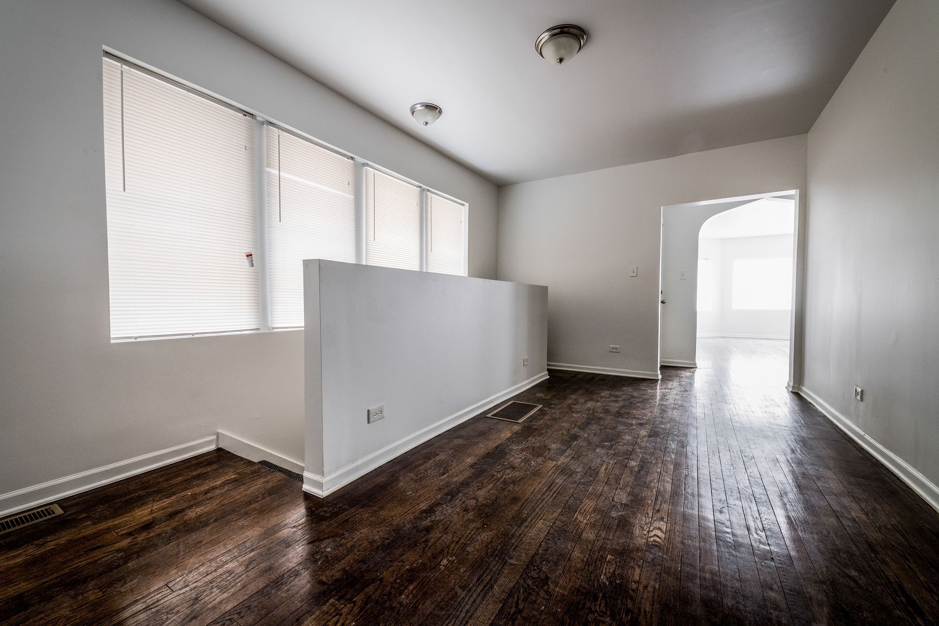 Empty room with wood floors, a partial white wall, windows with blinds, and doorway.