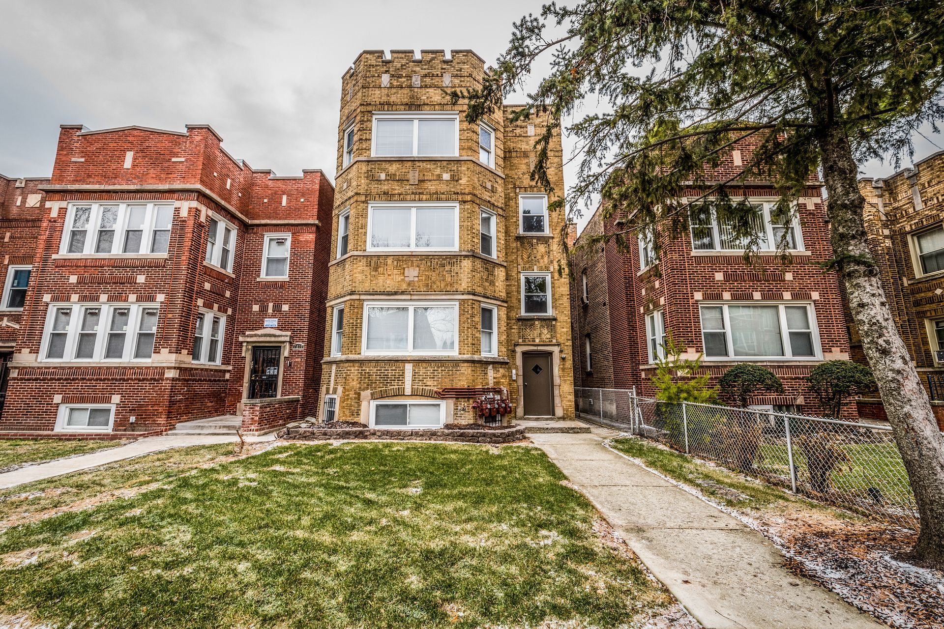 Row of three-story brick apartment buildings with green lawns and a cloudy sky.