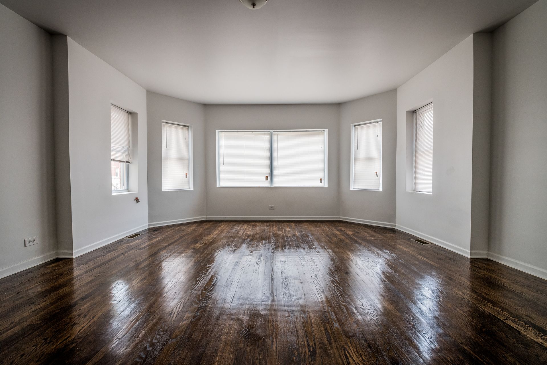 Empty room with dark hardwood floors, white walls, and a bay window with blinds.