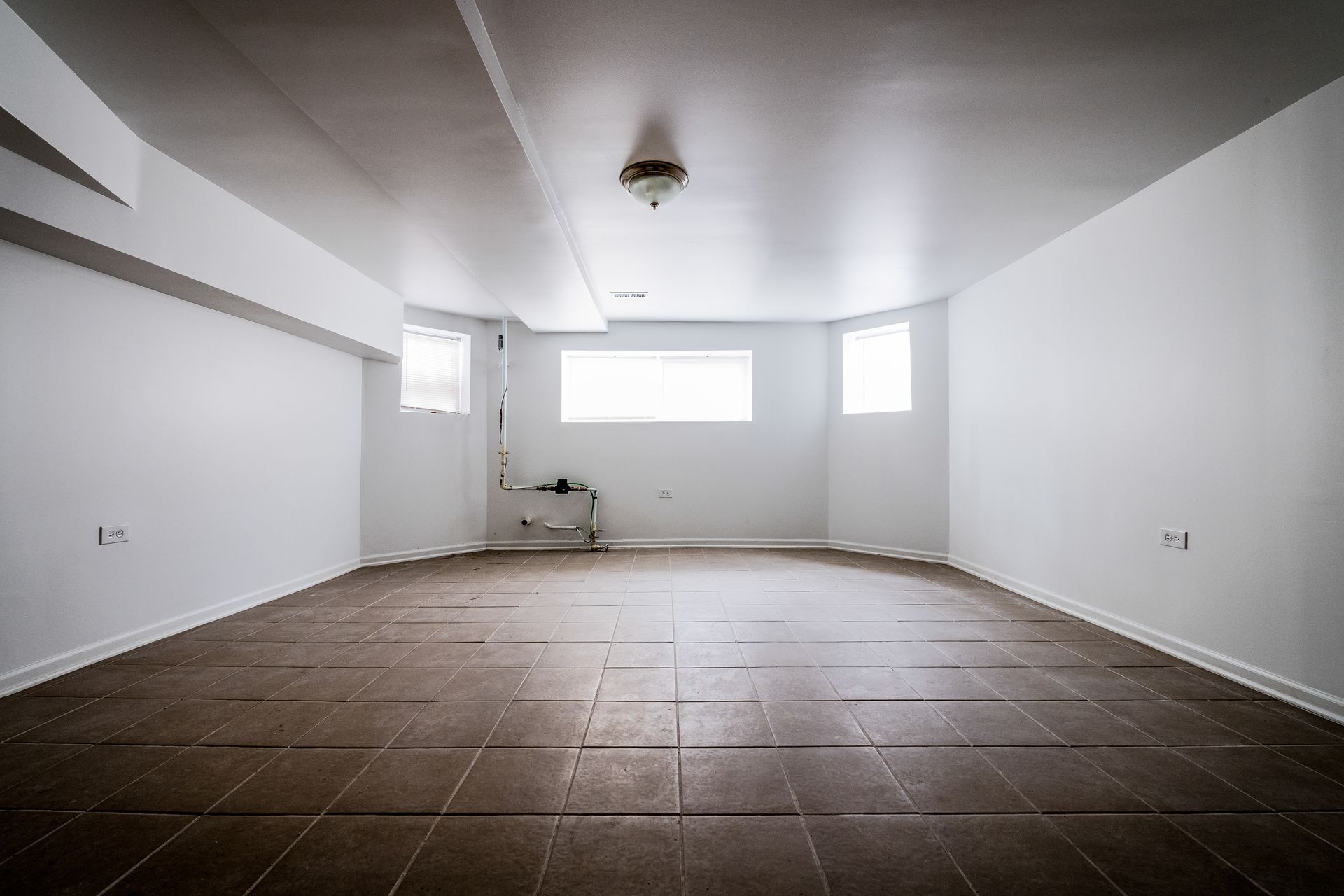 Empty basement room with white walls, brown tile floor, and a window.