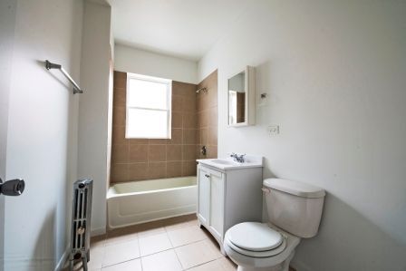 Bathroom with white walls, tan tile, window above tub, white toilet and vanity.