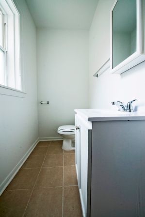 Small, neutral-toned bathroom with a sink, toilet, window, and mirror. Tan tiled floor, white walls and cabinet.
