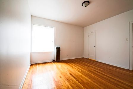 Empty room with hardwood floors, white walls, window, radiator, and a closed door.