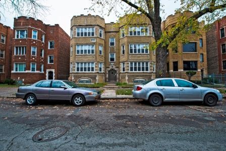 Cars parked on street in front of brick apartment buildings with mature tree.