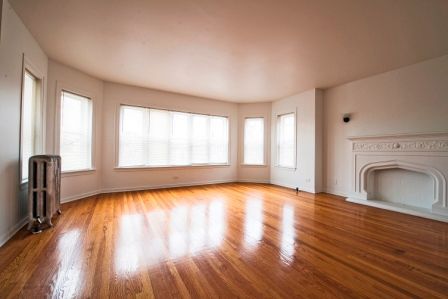 Empty living room with hardwood floors, white walls, and a fireplace. Large windows provide natural light.