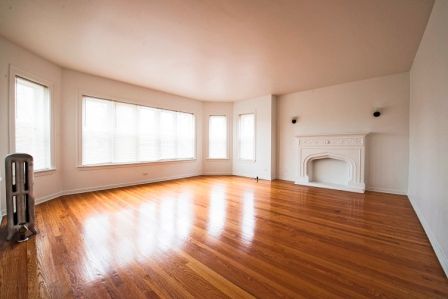 Empty living room with hardwood floors, white walls, and fireplace. Large window with sheer curtains.