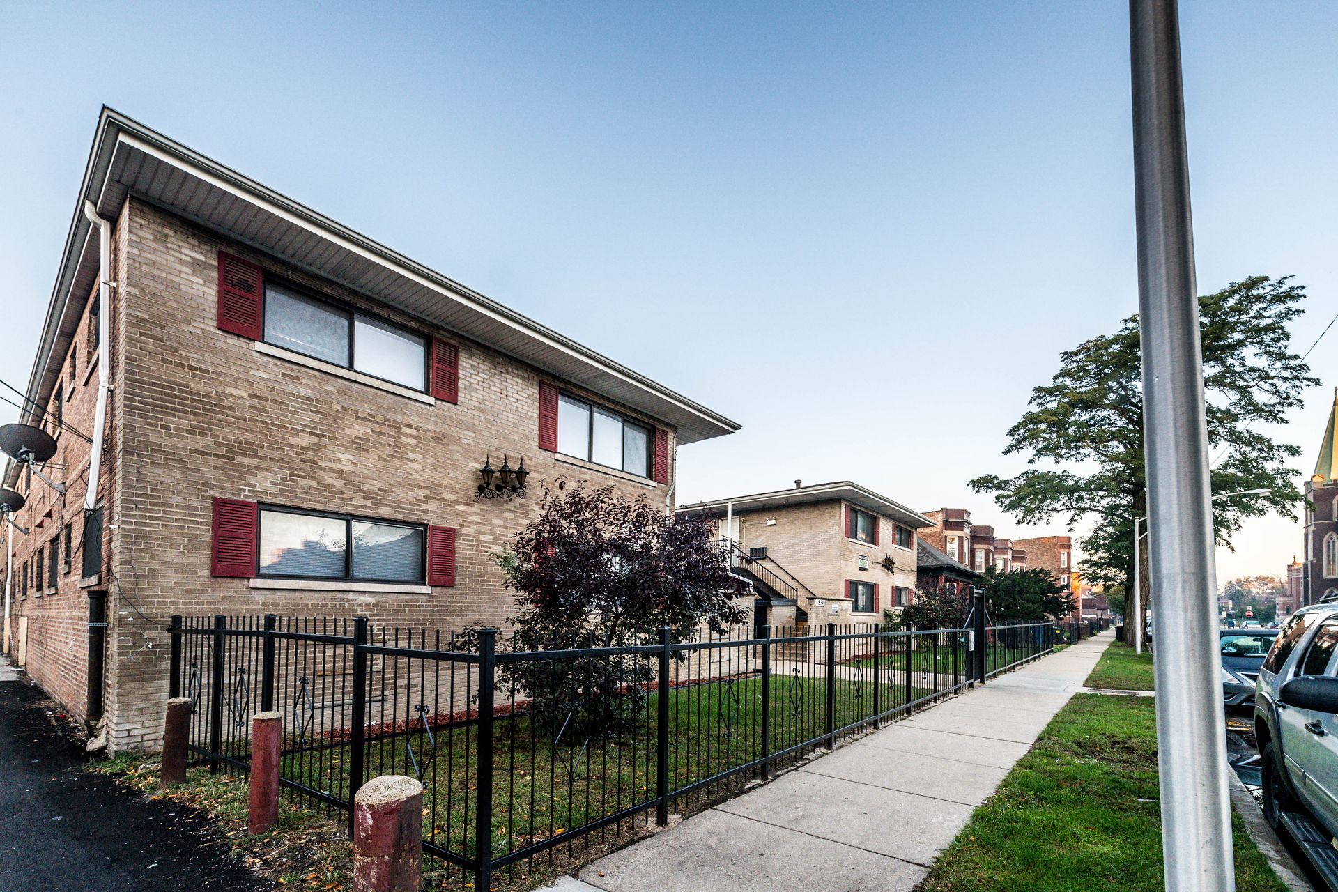 Two-story brick apartment building with red shutters and black fence along a sidewalk, street view.