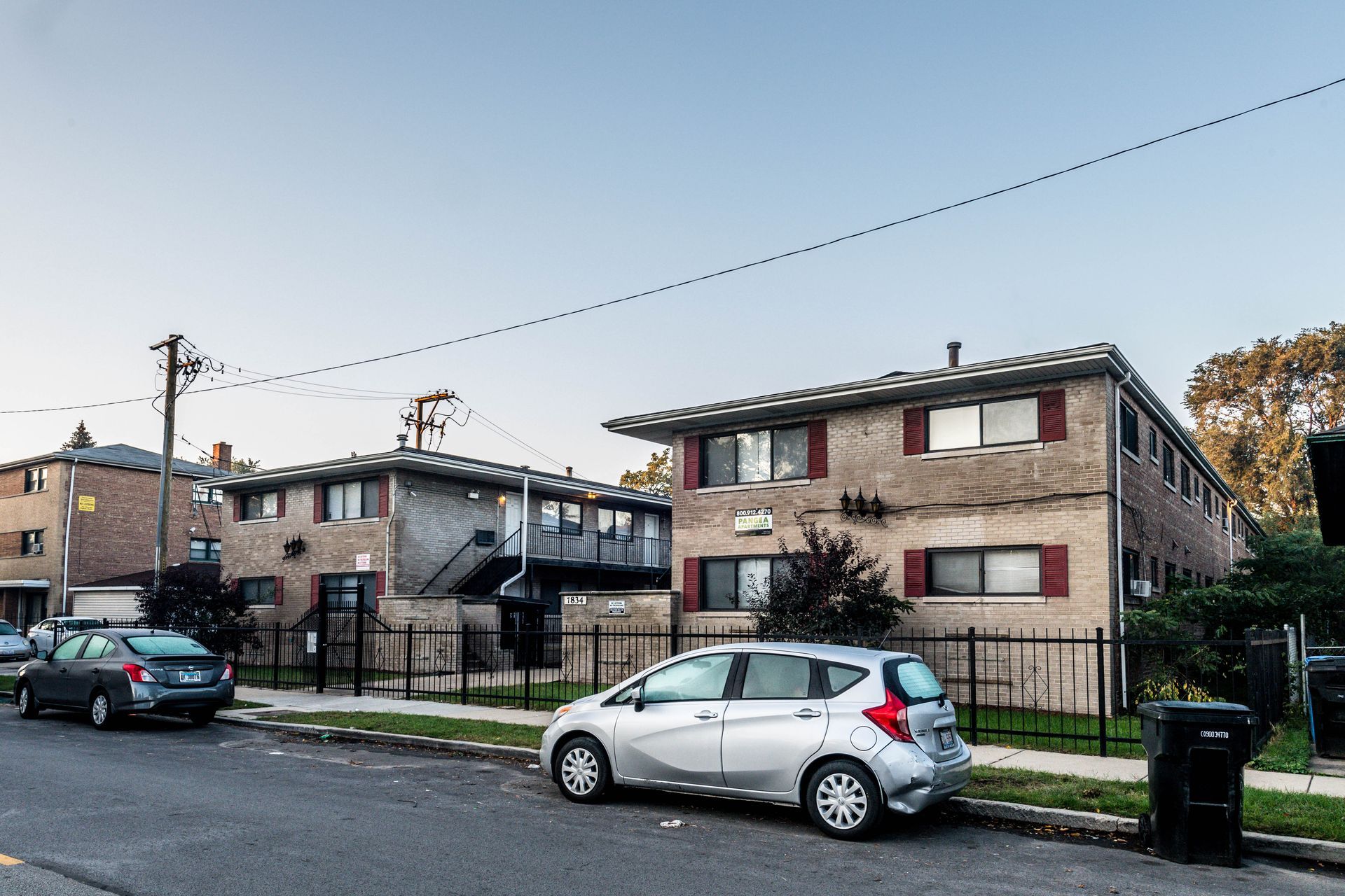 Two-story brick apartment buildings with cars parked on the street, behind a black metal fence, on a clear day.