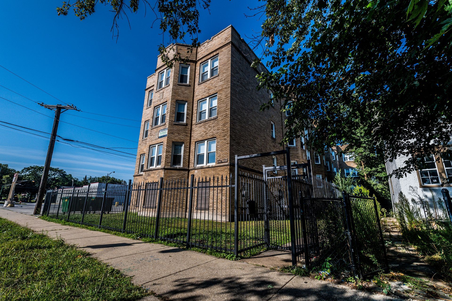 Brick apartment building behind a black metal fence on a sunny street, trees and sidewalk in view.