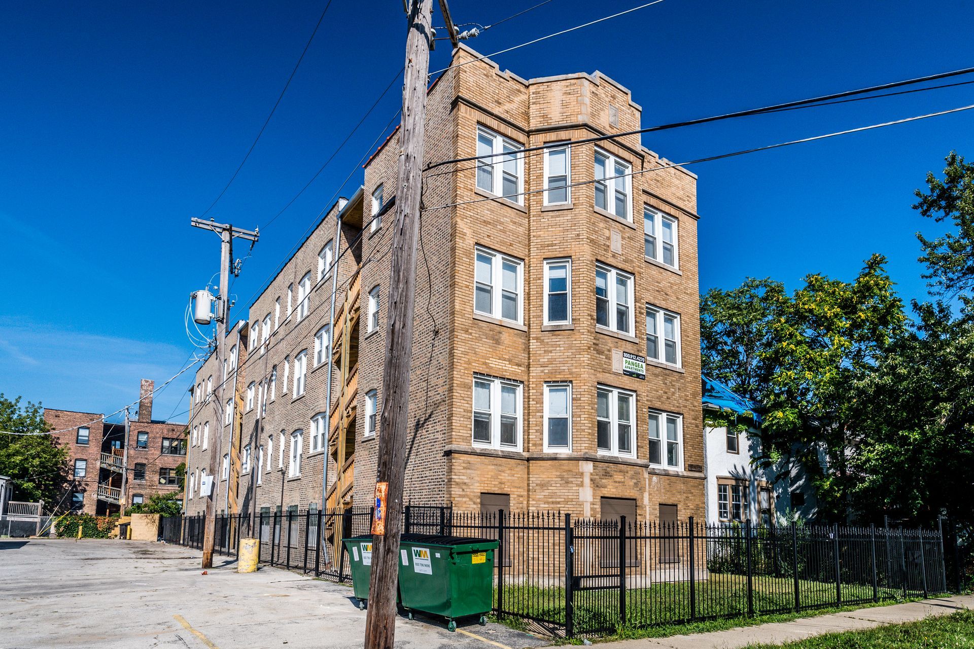 Multi-story brick apartment building on a sunny street, green dumpster, utility poles, trees, and blue sky.