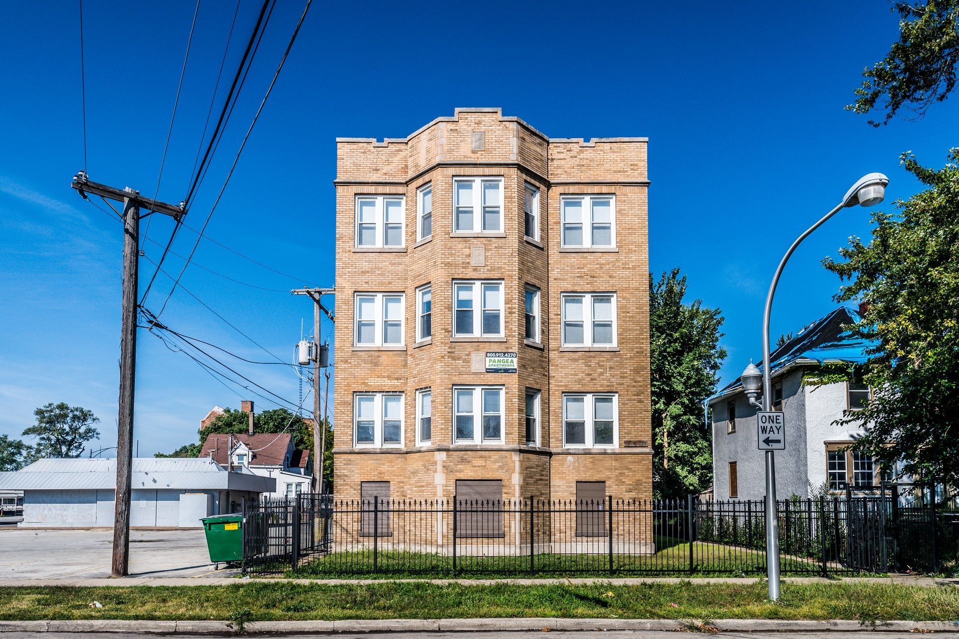 Four-story tan brick apartment building with iron fence, power lines, and blue sky.