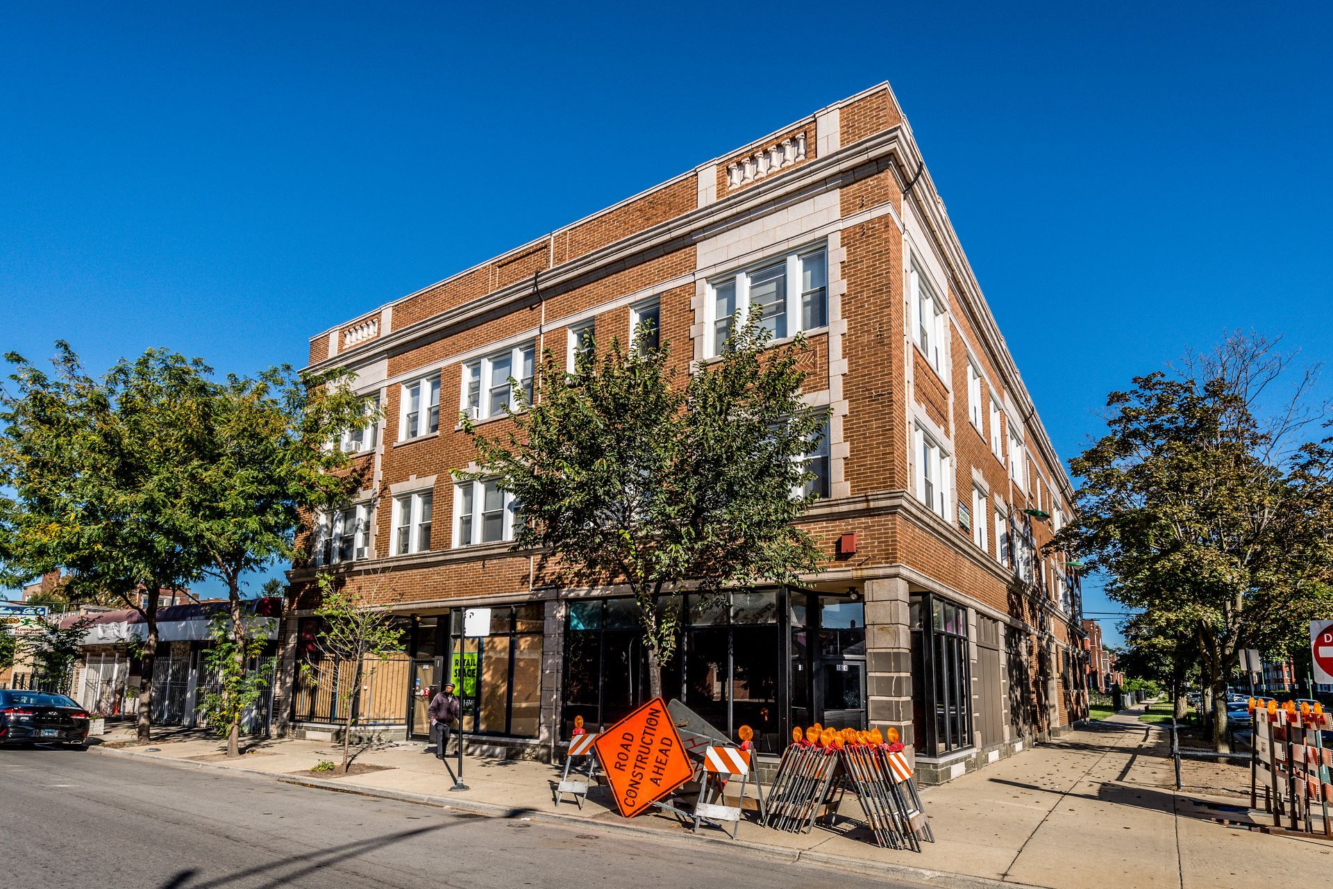 Three-story brick building on a sunny day with storefronts below, a construction area in front, and trees on the sidewalk.