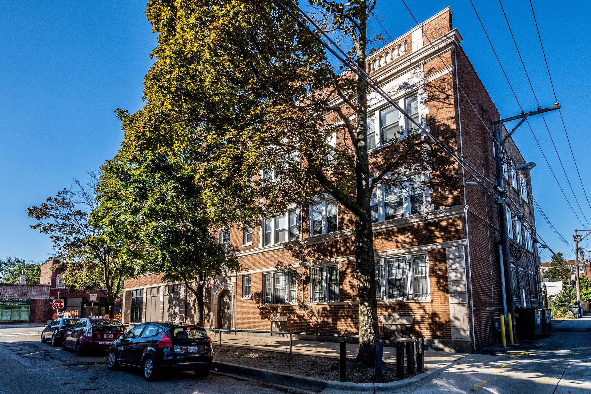 Brick apartment building on a street with parked cars and trees; sunny day.