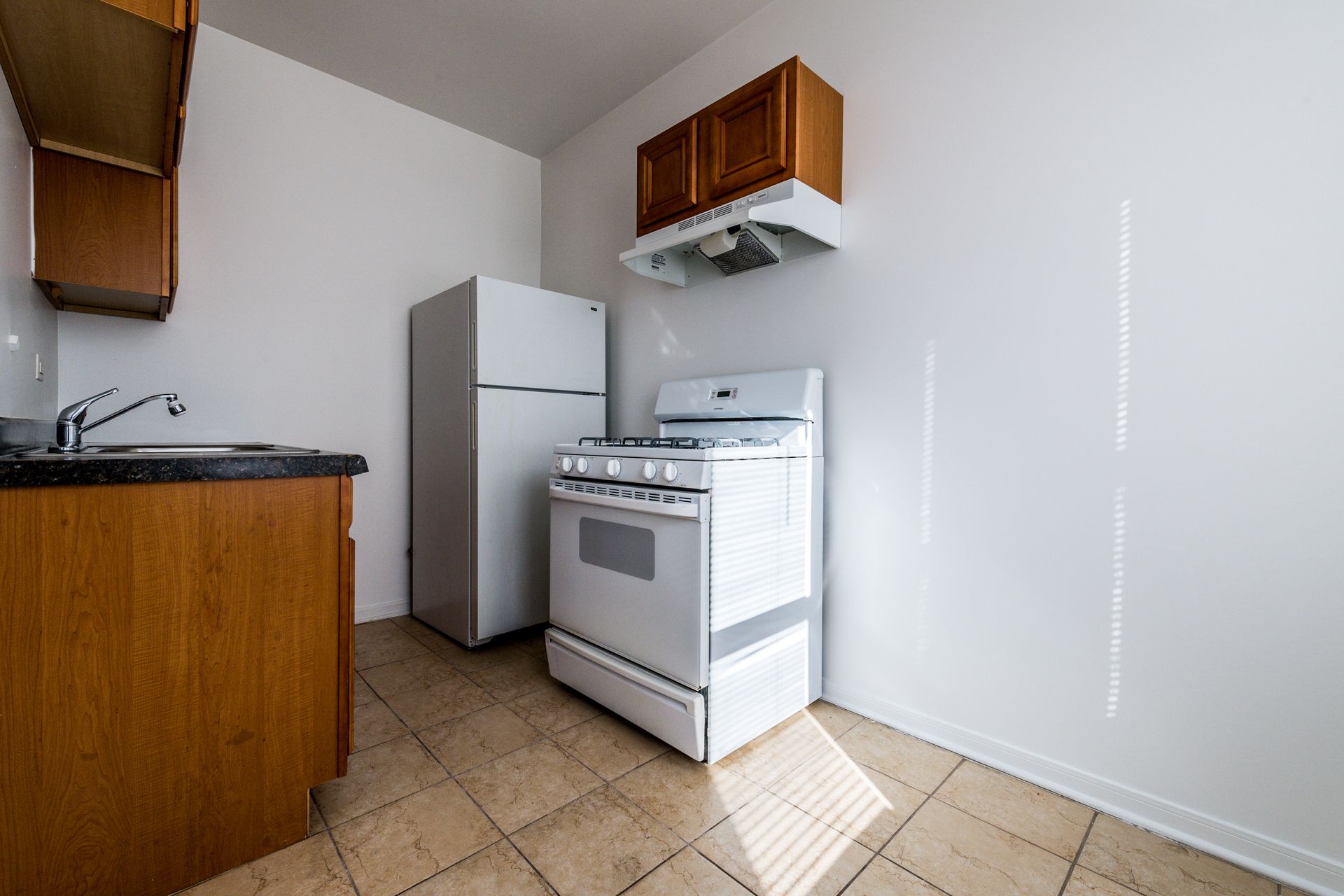 Small kitchen with white appliances, wooden cabinets, and tan tile floor.