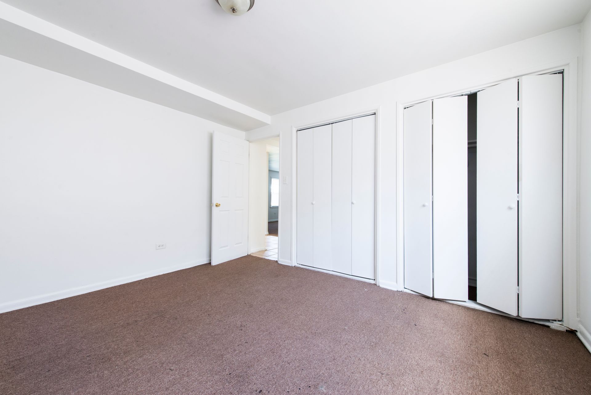 Empty bedroom with white walls, brown carpet, and closet doors.