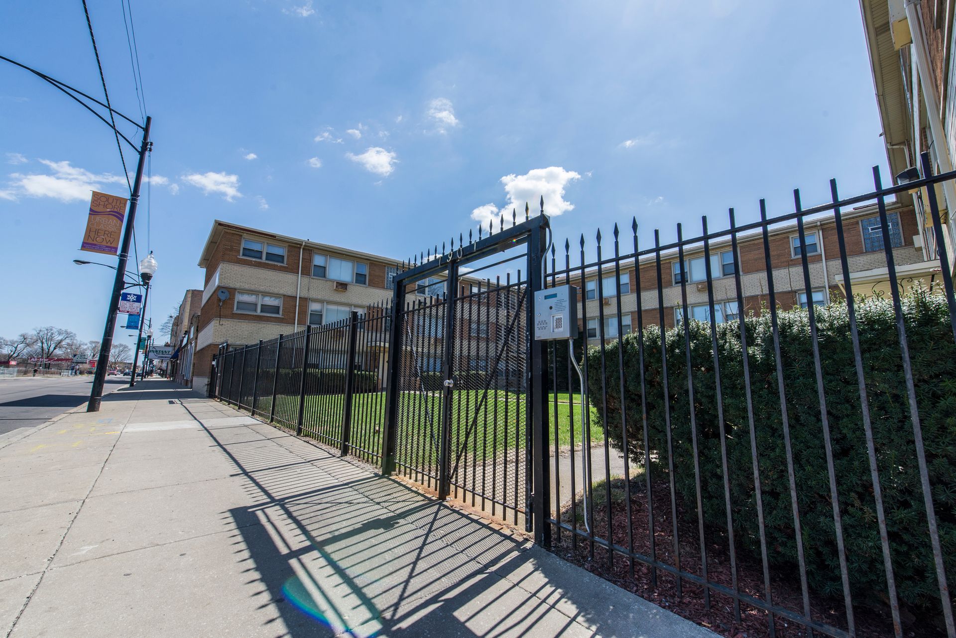 Black metal fence encloses a grassy area in front of a brick apartment building; a sidewalk and street are visible.