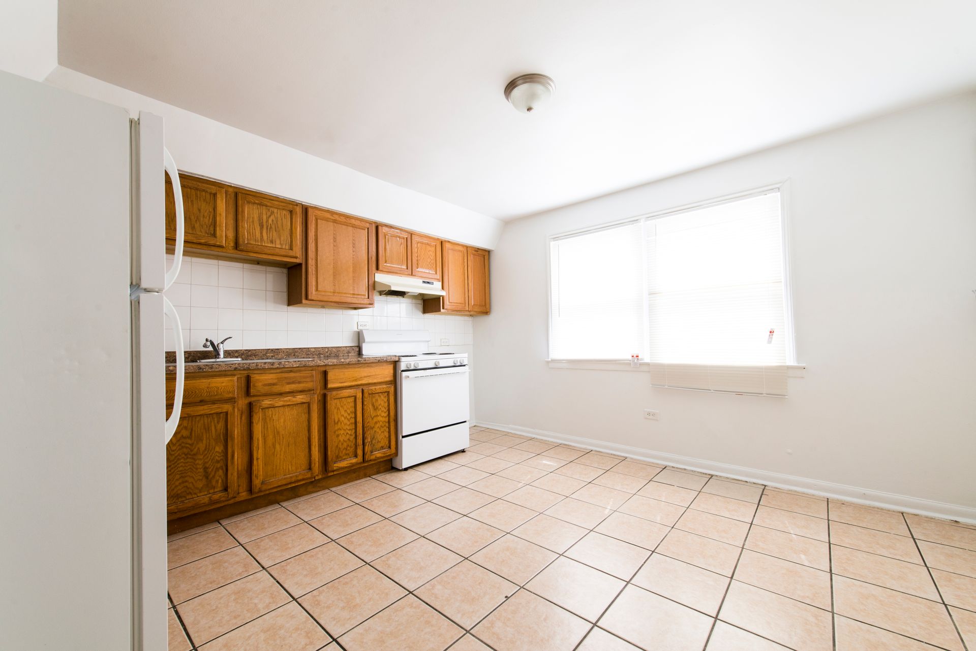 Kitchen with wooden cabinets, white appliances, and tiled floor. Light streams in from the window.