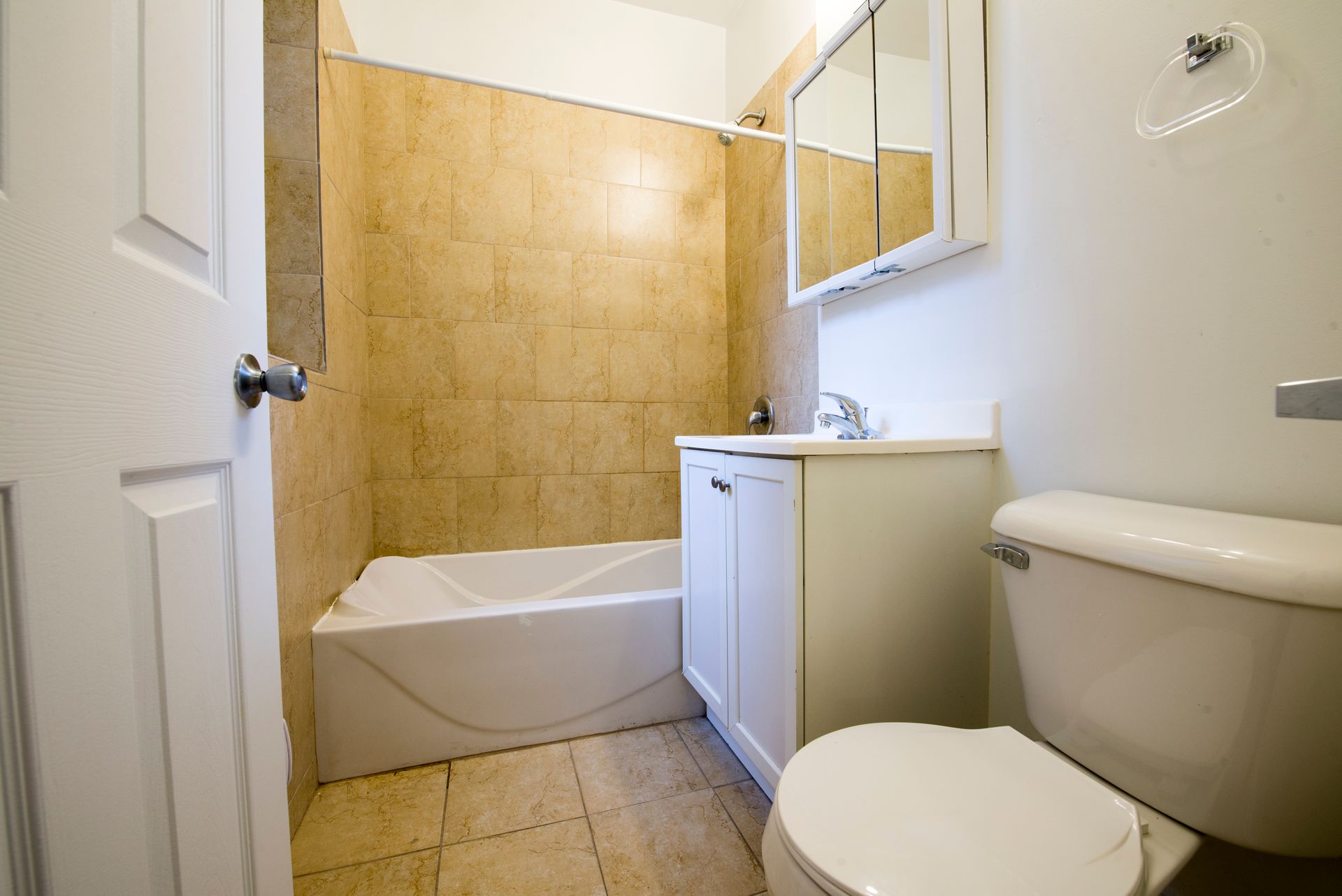 Bathroom with a white toilet, vanity, and bathtub; tan tiled walls, and a mirrored cabinet.