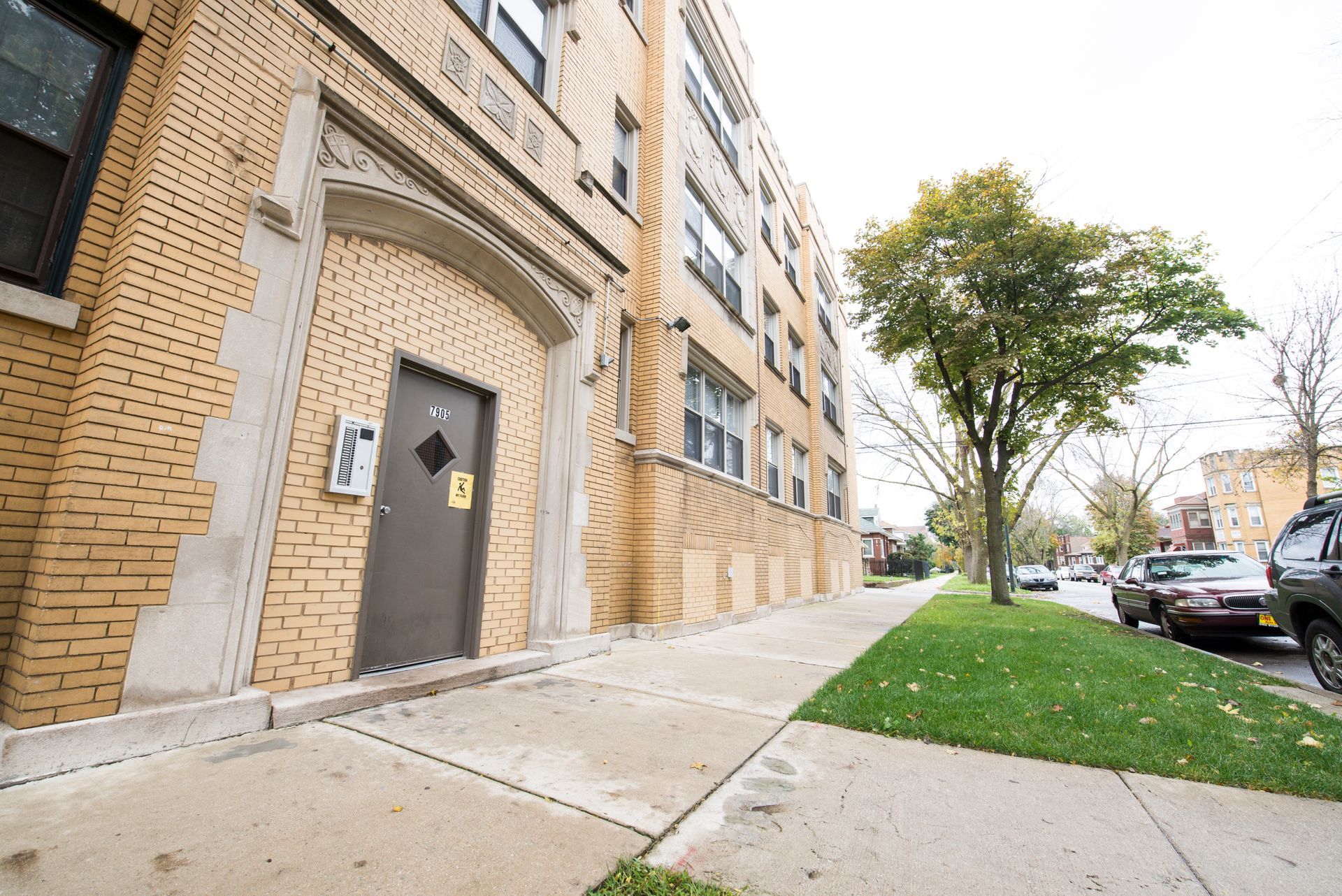 Brick apartment building with sidewalk, street, cars, and a tree.