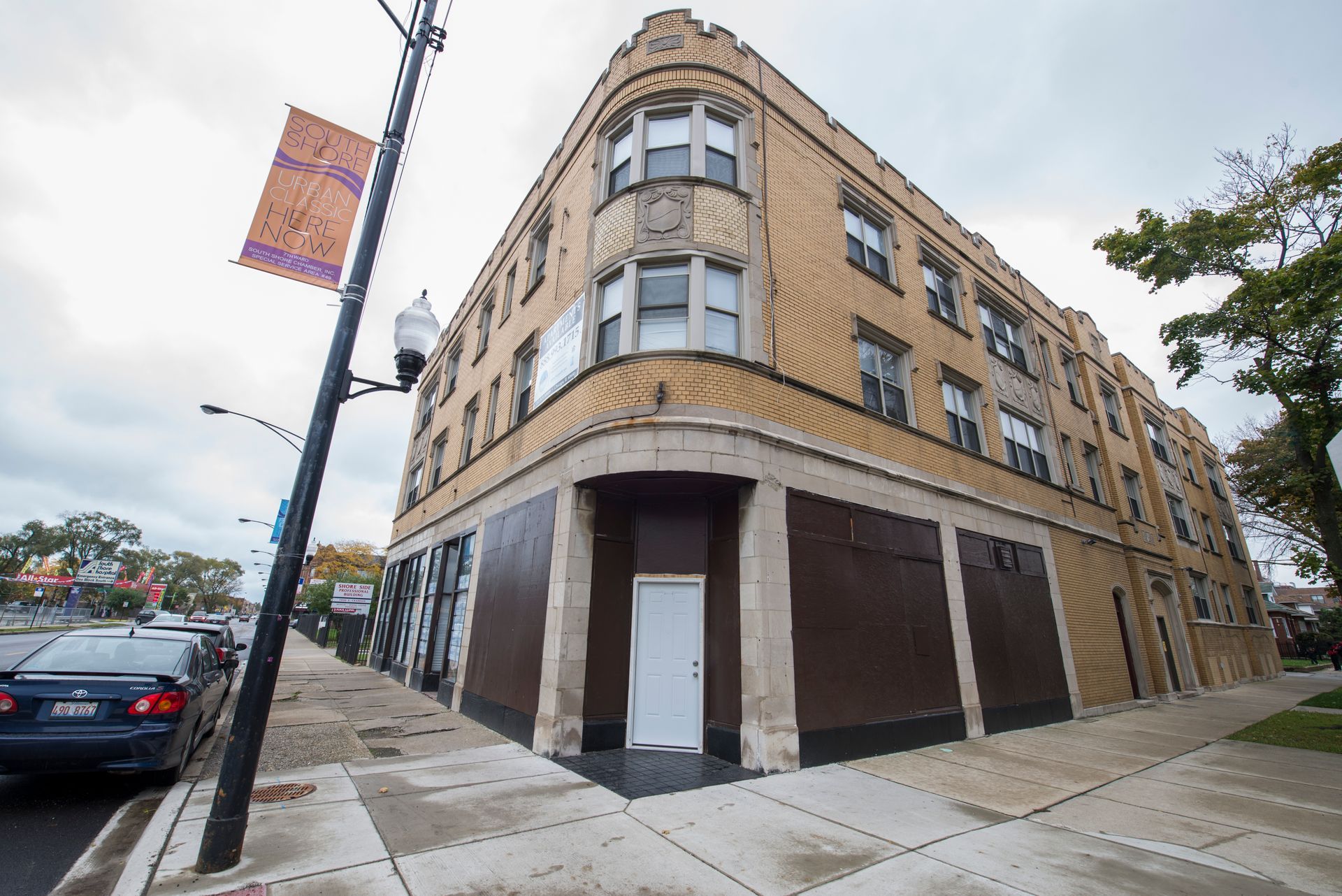 Corner apartment building with boarded-up storefronts, a banner, and a street with parked cars.