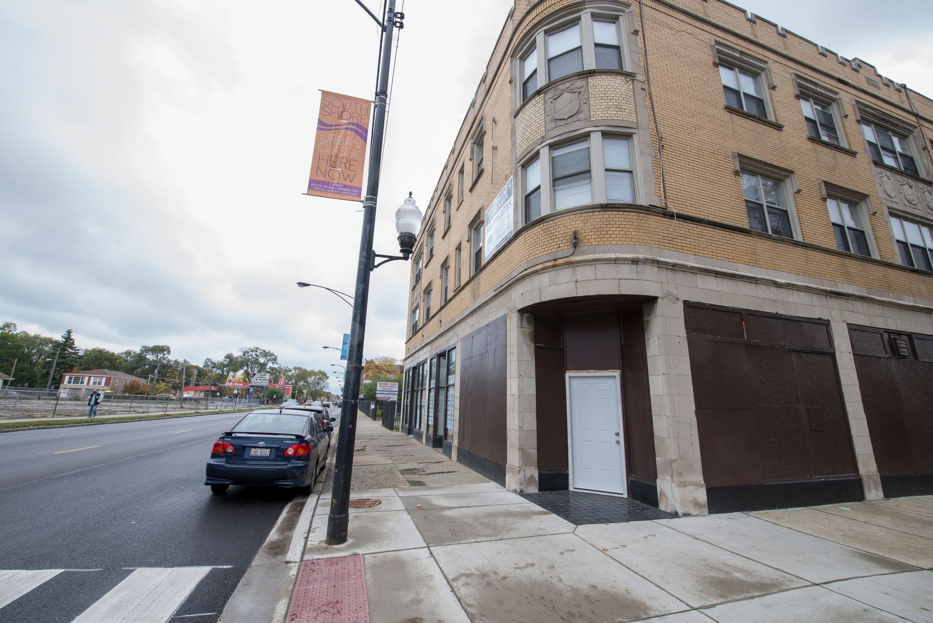 A car parked near a building with boarded-up storefronts and a banner on a cloudy day.