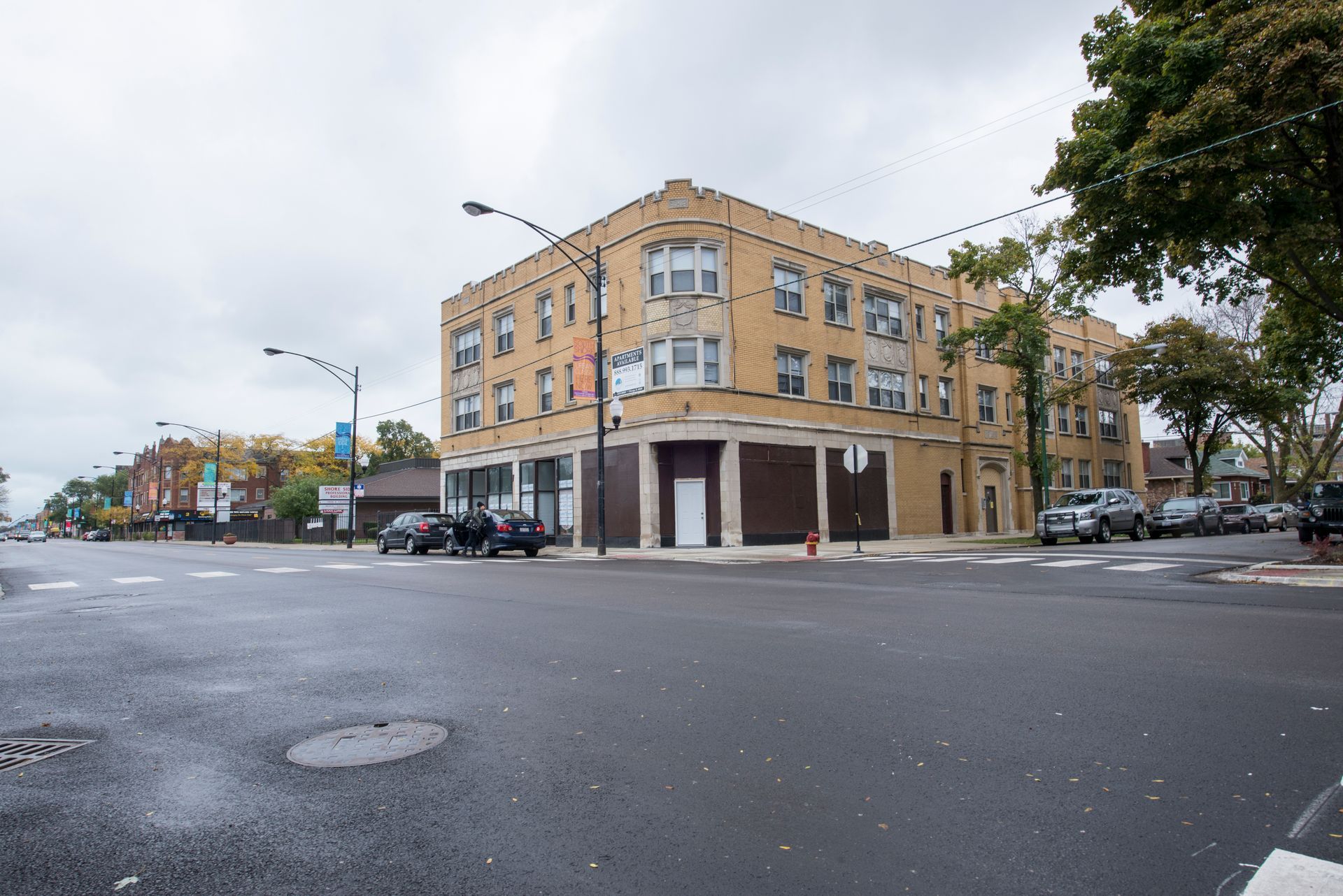 Corner building with boarded-up storefronts and residential units above; cloudy sky, street view.