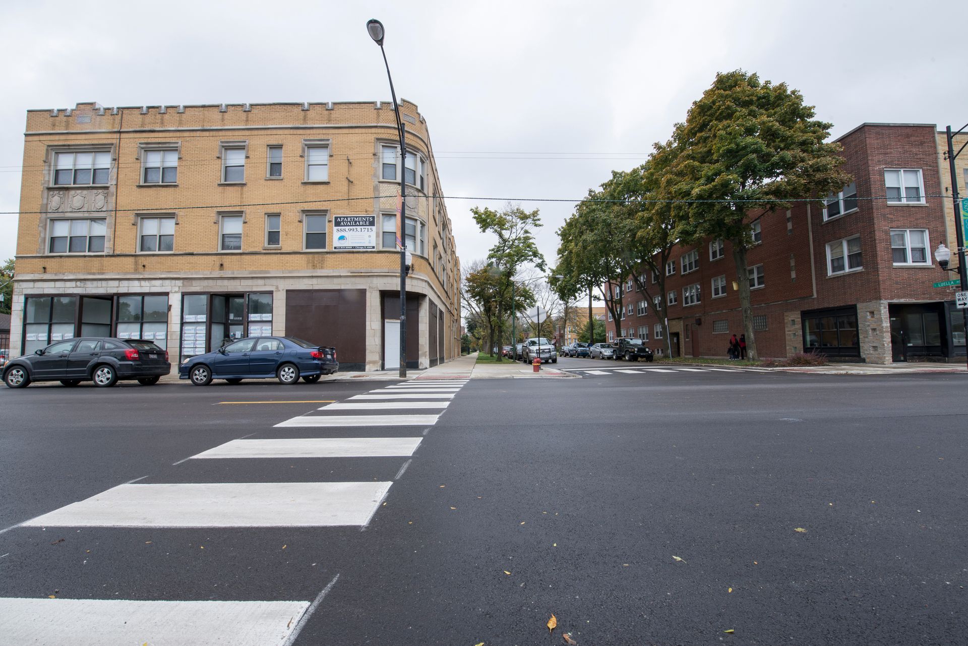 Street scene with crosswalk, buildings, parked cars, and overcast sky.