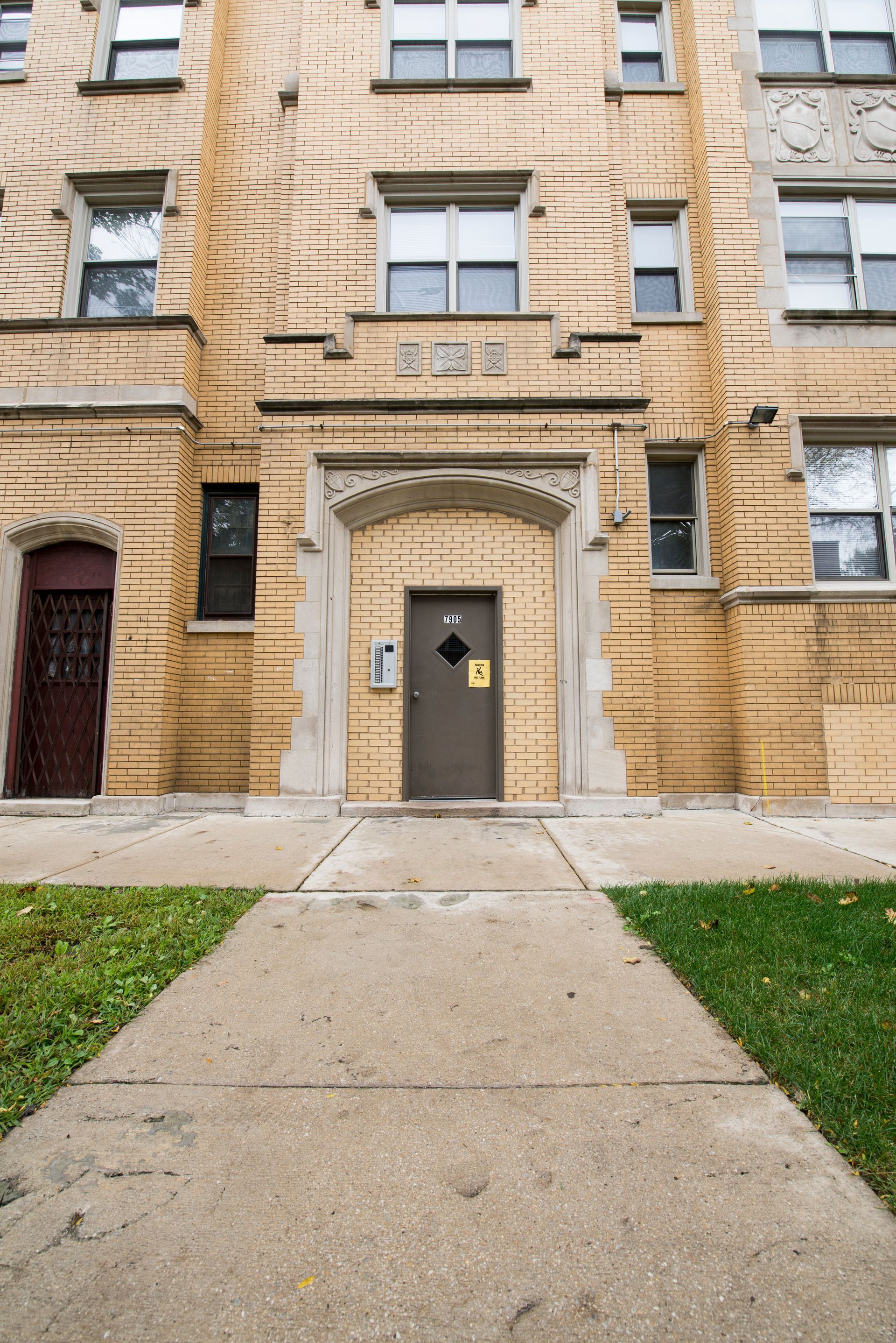 Building entrance with brown door, arched doorway, and tan brick facade. Concrete path leads to the door.