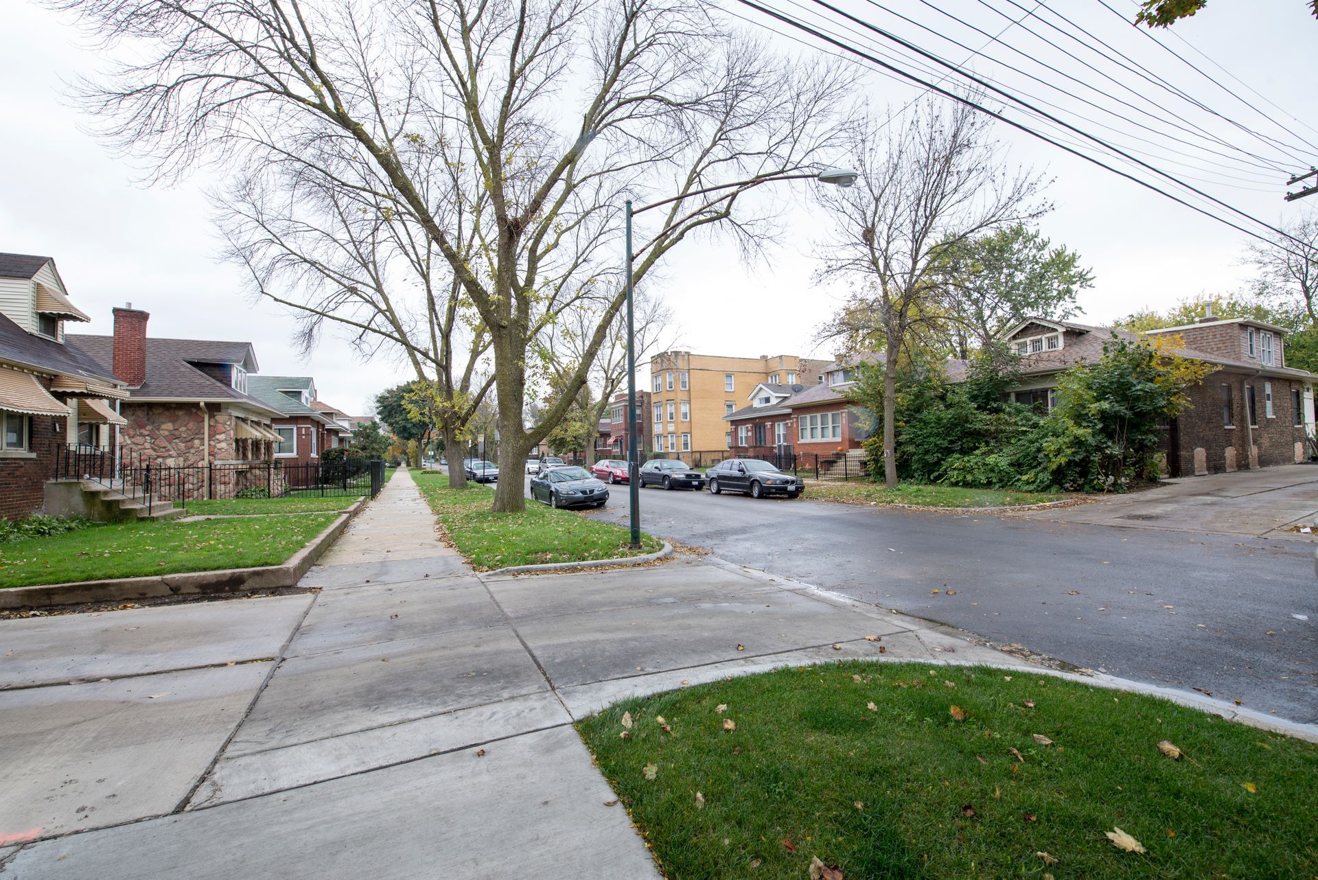 Sidewalk view of a residential street with parked cars and houses on a cloudy day.