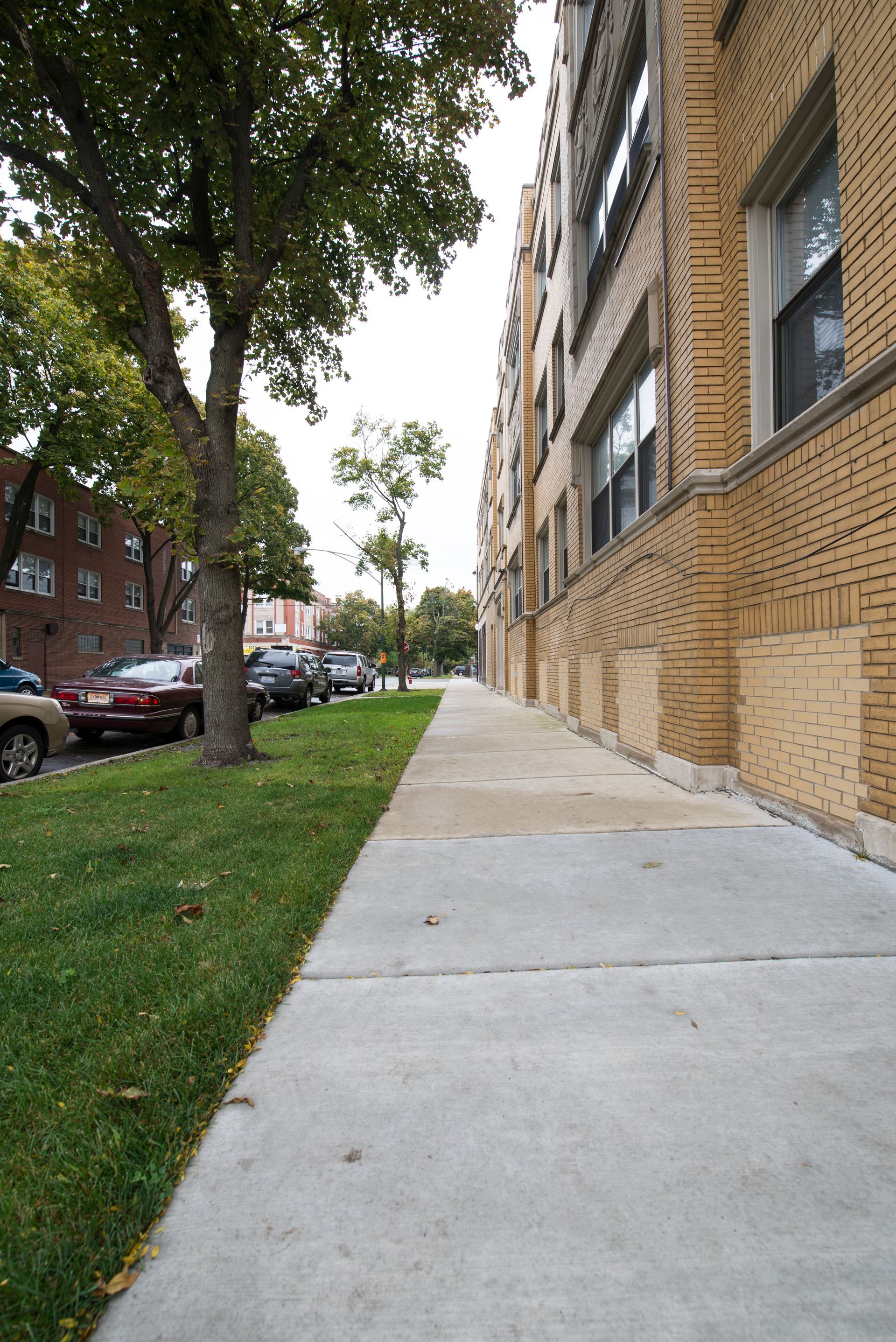 Sidewalk along a brick apartment building with trees and parked cars.