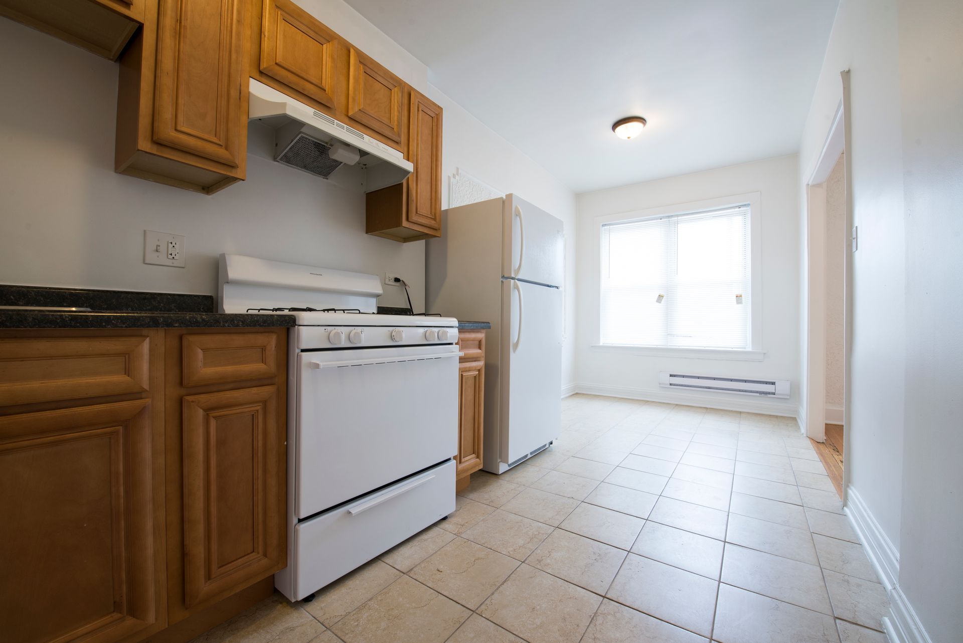Kitchen with wooden cabinets, white appliances, and tiled floor. Light streams in from a window.