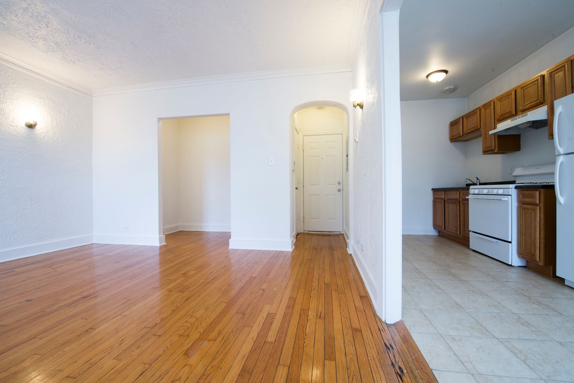 Empty apartment interior with hardwood floors, white walls, and kitchen visible.