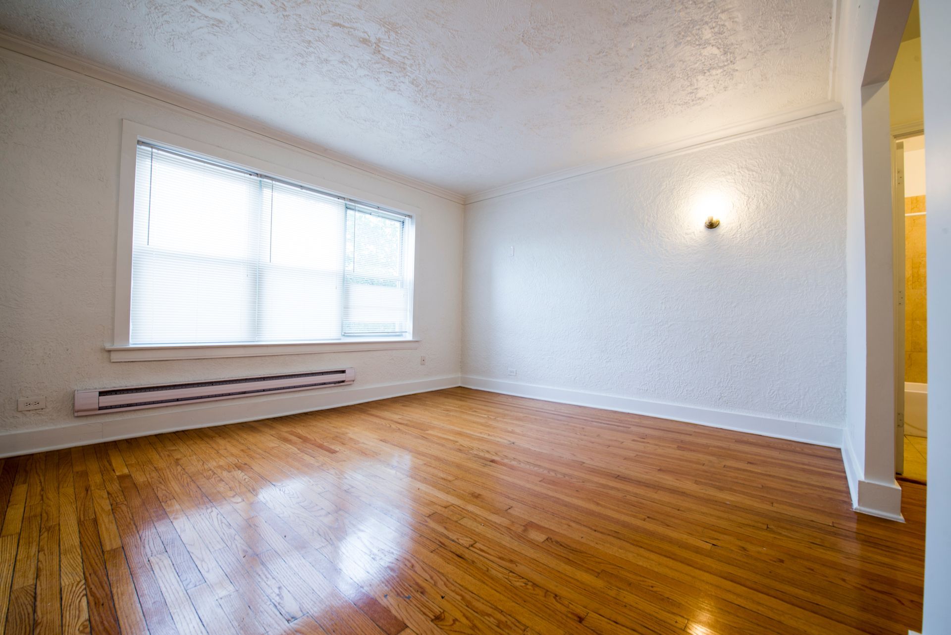 Empty room with hardwood floors, a large window with blinds, and white walls.