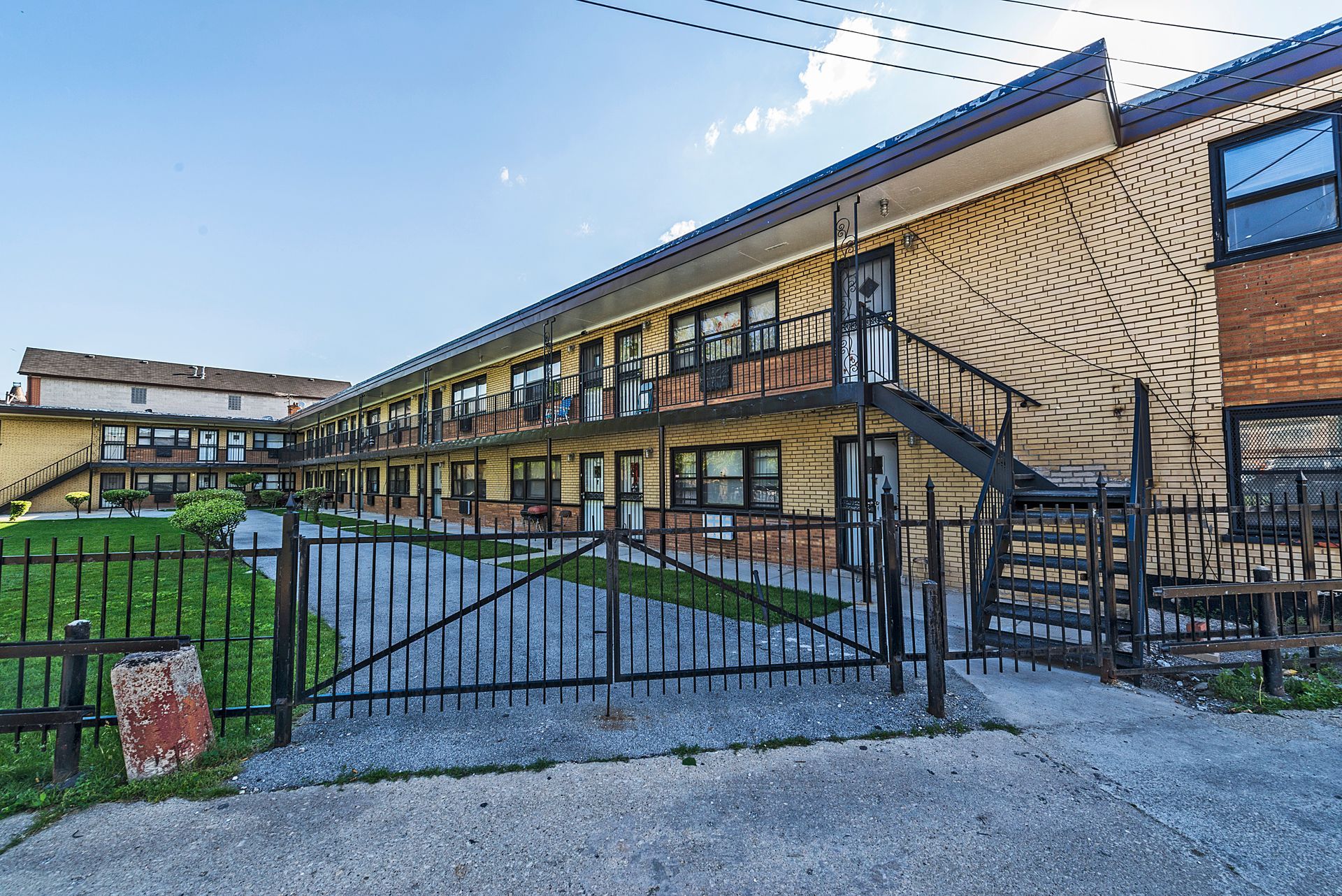Two-story brick apartment building with black metal staircases, a gate, and a green lawn.