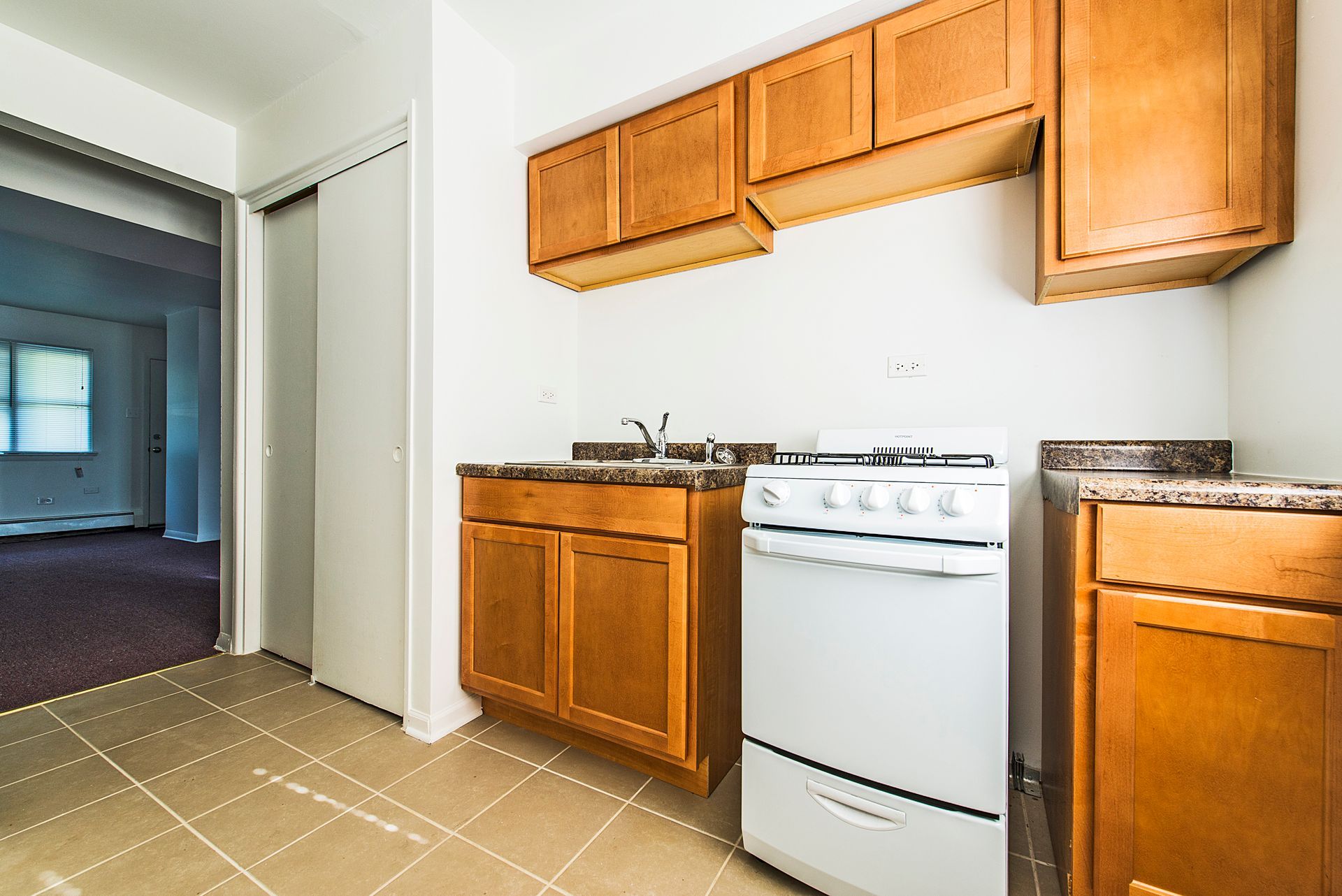 Small kitchen with wooden cabinets, white stove, and stone countertops; doorway to another room.