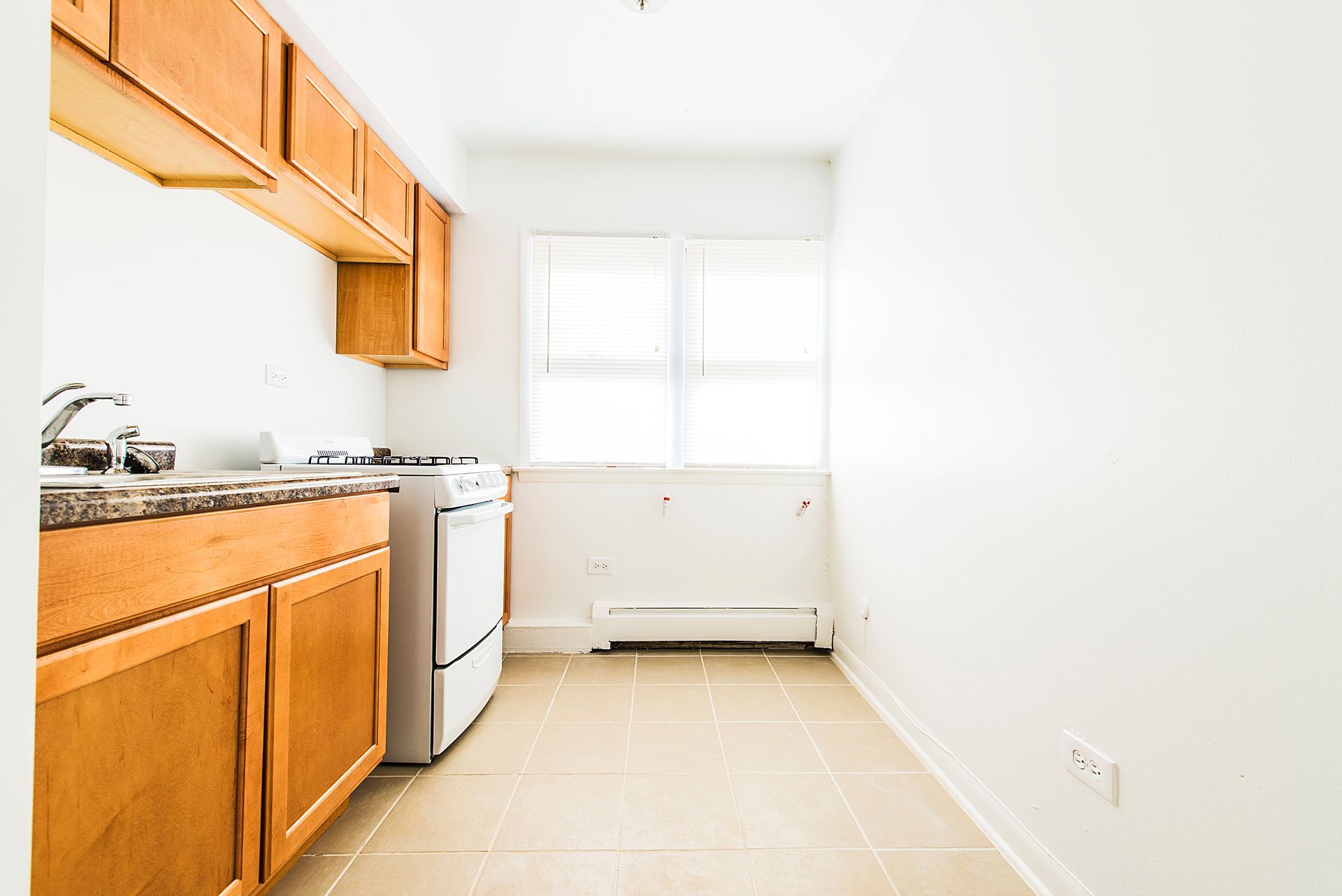 Small kitchen with light brown cabinets, white stove, and window with blinds.