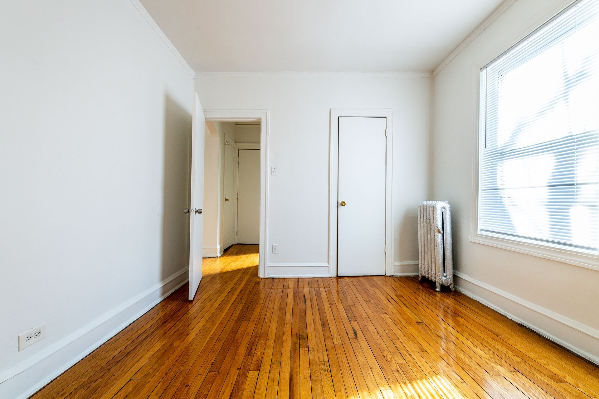 Empty room with hardwood floors, white walls, two doors, and a window with blinds.