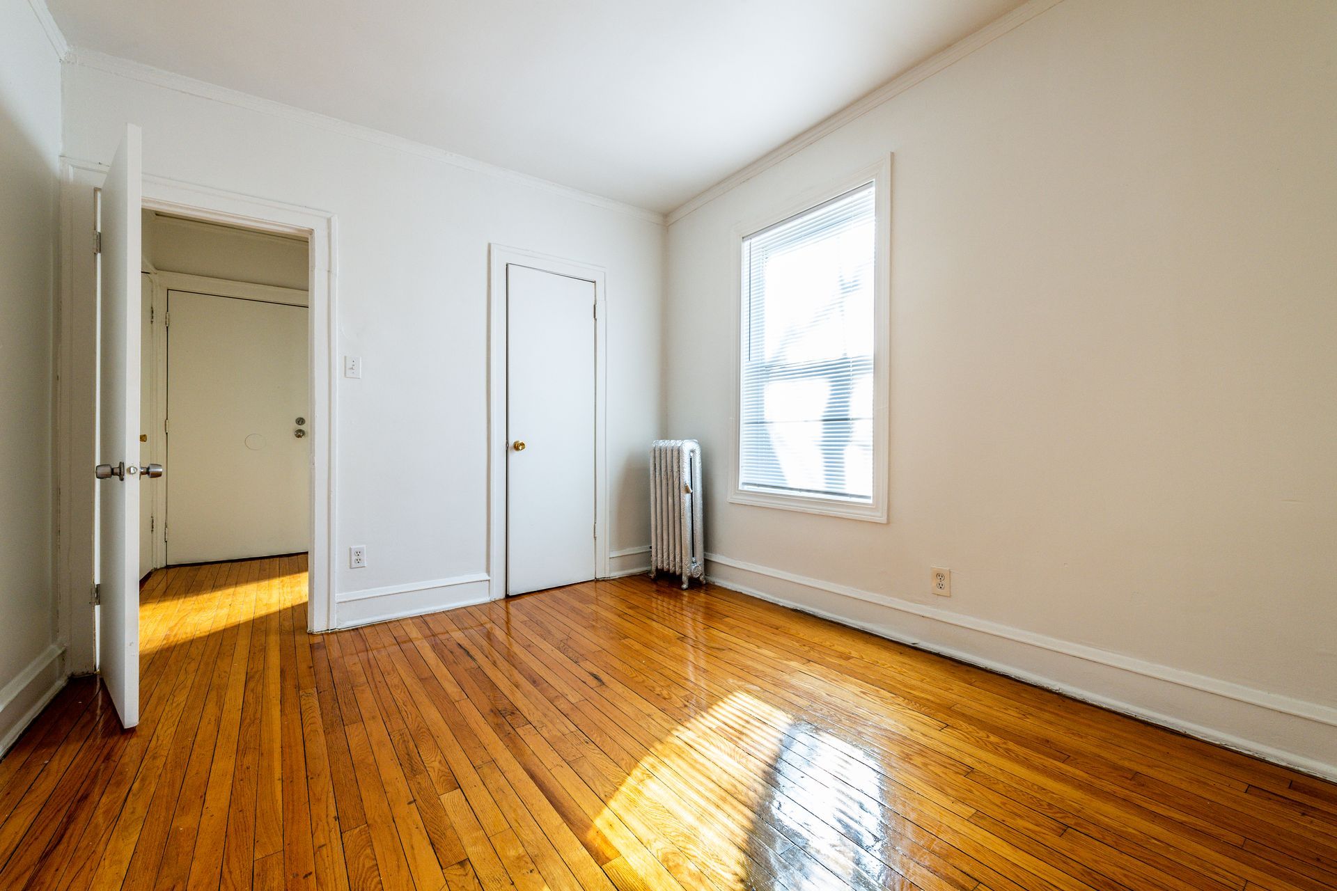 Empty room with hardwood floors, white walls, closed door, and window with blinds.