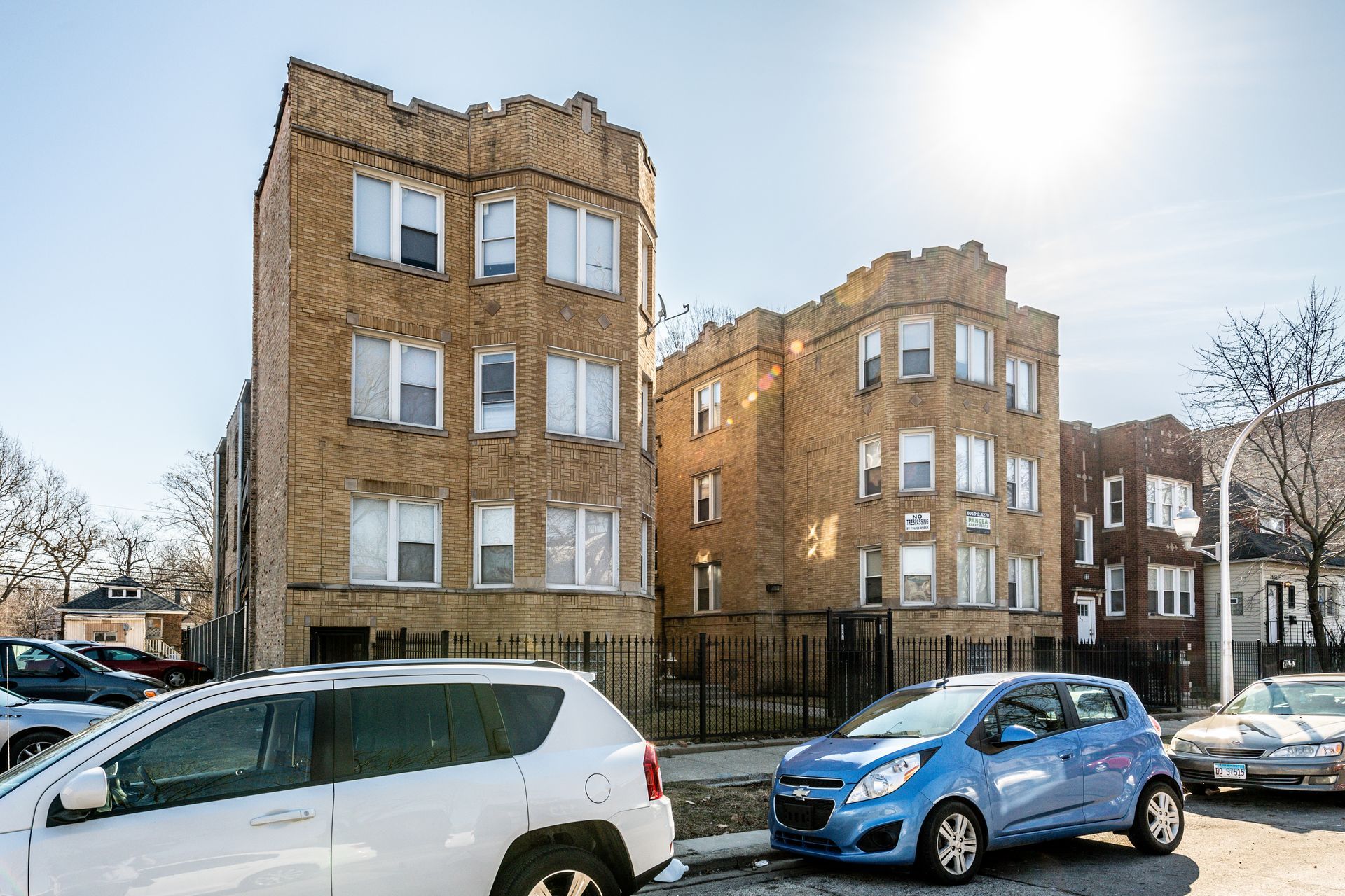Two brick apartment buildings with cars parked on the street on a sunny day.