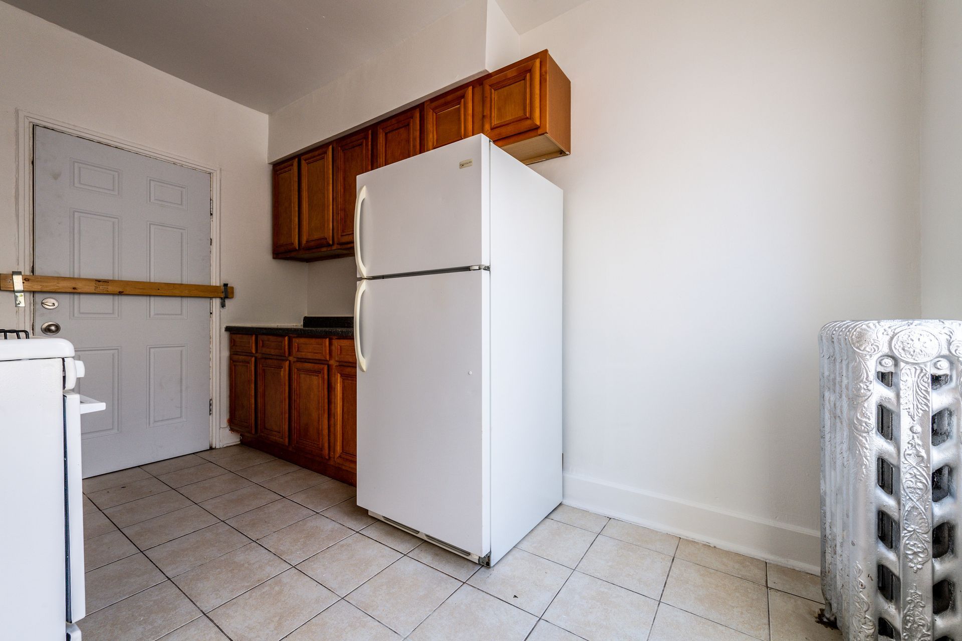 Kitchen with white fridge, wooden cabinets, and tile floor. A white door is on the left.
