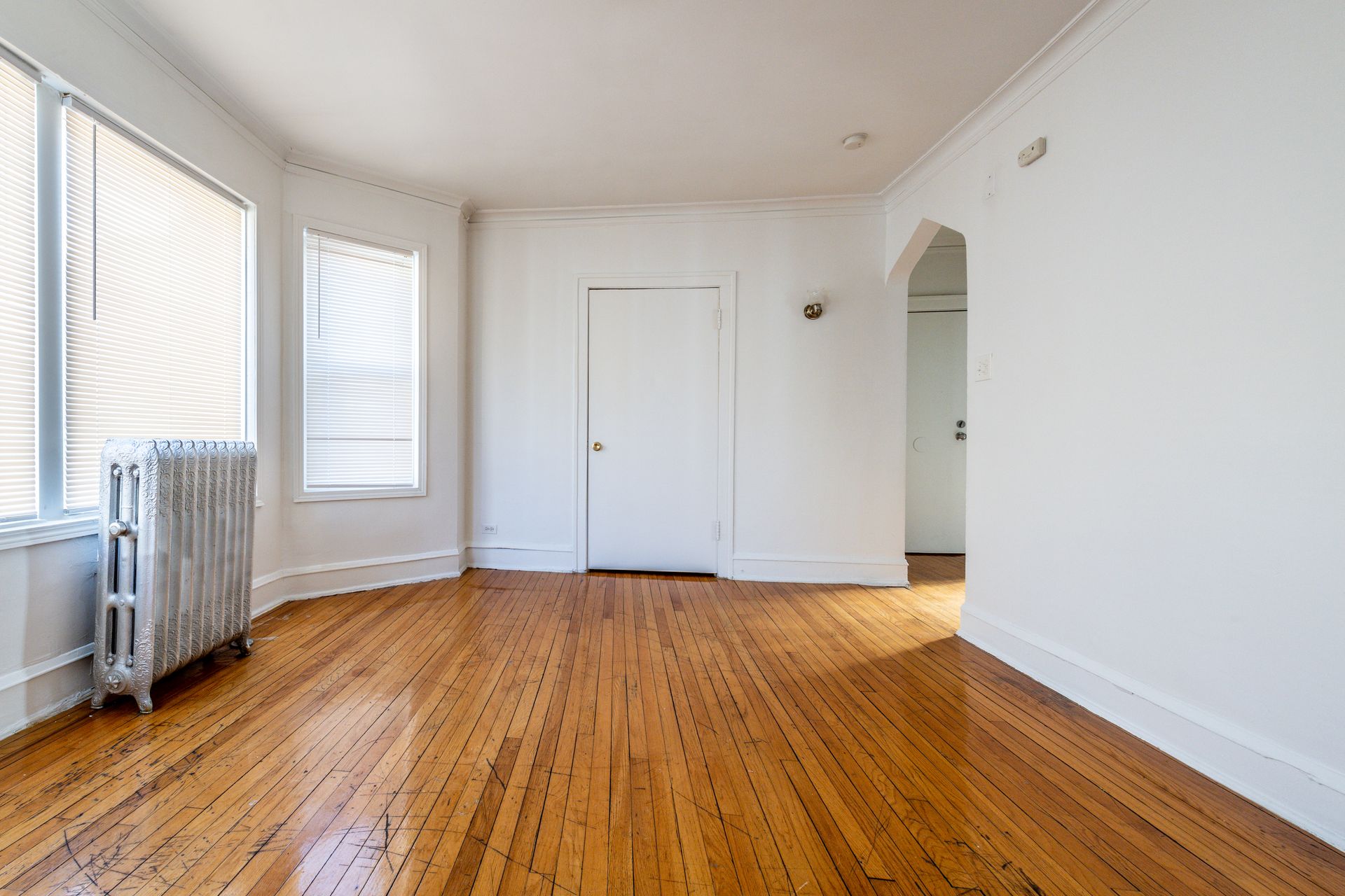 Empty room with hardwood floors, white walls, large windows, radiator, and arched doorway.